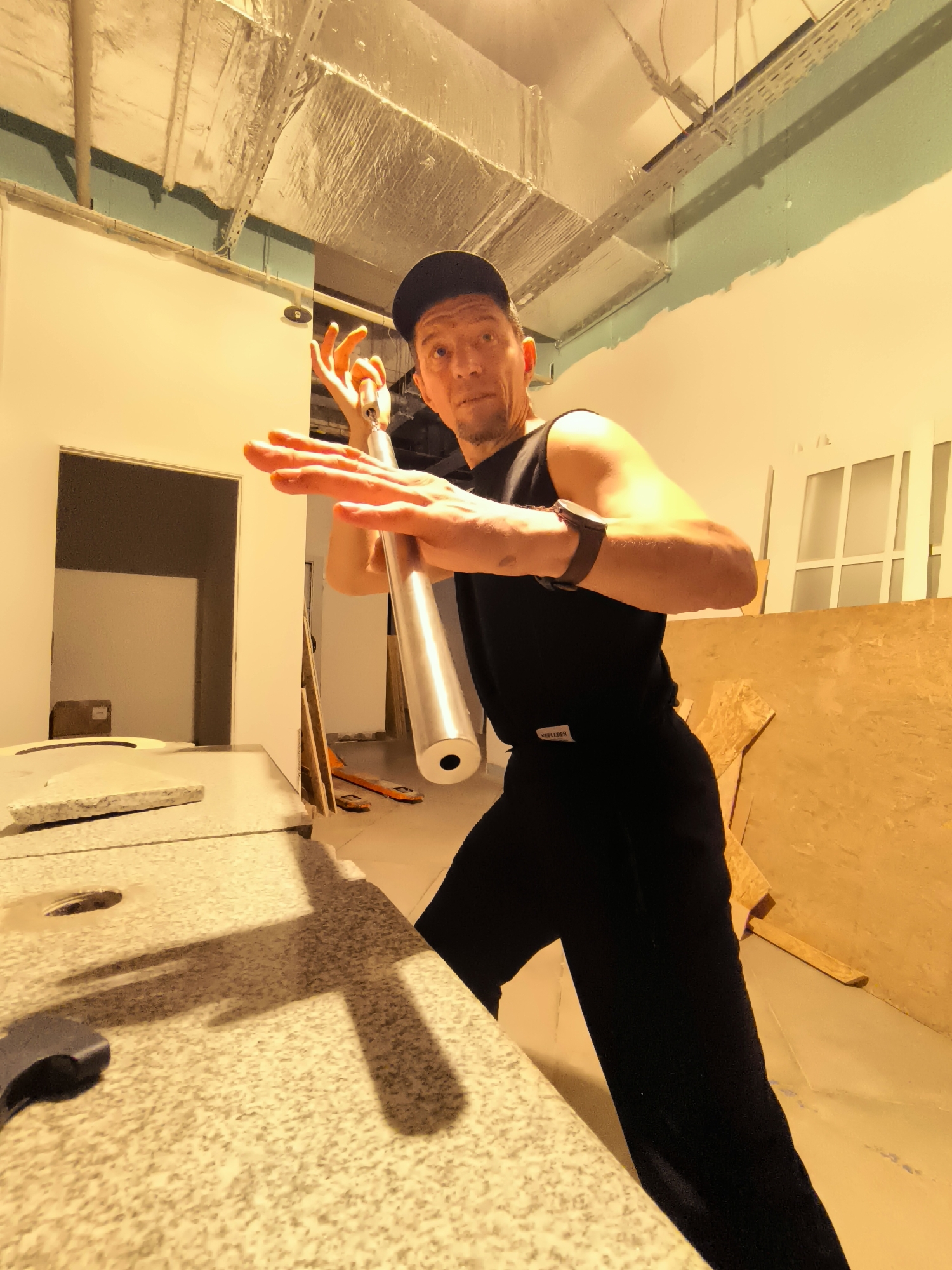 A man in a black baseball cap and black sleeveless shirt is posing in an unfinished room. He is holding a silver cylindrical object, possibly a piece of pipe, in front of him with both hands, his right hand on top and his left hand below. His left arm is bent at the elbow, and his right arm is extended. His fingers are spread wide in what appears to be a martial arts pose. He is wearing black pants and a smartwatch on his left wrist. The room has exposed ceiling pipes and insulation, light-colored walls, and some construction materials, including large sheets of plywood leaning against a wall. A doorway is visible in the background. The flooring appears to be speckled, possibly granite or a similar material, and there is a dark-colored tool on the floor in the foreground, casting a shadow. The lighting in the room is warm and casts long shadows. This image does not depict the city of Qarağandı city administration, Kazakhstan.