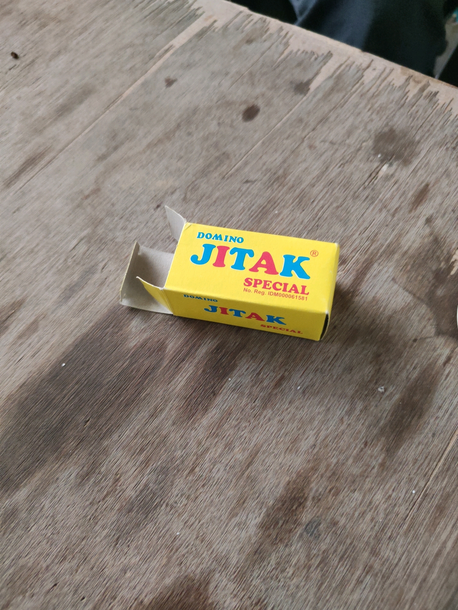 An open, yellow cardboard box with the brand name "DOMINO JITAK SPECIAL" printed in blue and red lettering sits on a wooden surface. The box is slightly ajar, with one flap open. The wood grain of the surface is visible, with various shades of brown and grey. The setting appears to be outdoors or in a rustic indoor environment. No people are present in the image. The scene is located in Maburai, Indonesia.