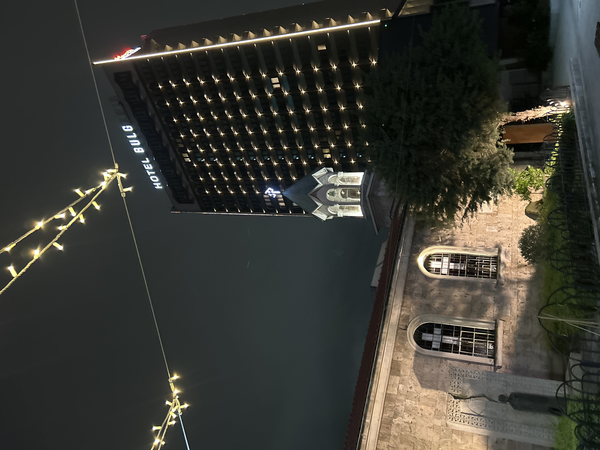 The image shows a nighttime scene in Burgas, Bulgaria, featuring the Hotel Bulgaria. The hotel is a high-rise building illuminated with numerous lights, and its name is displayed vertically in bright letters. Adjacent to the hotel, there's an older, lower building, potentially a church or historical site, characterized by stone architecture and arched windows. A string of festive lights adds to the scene's ambience. The sky is dark, indicating nighttime, and the overall impression is of a contrast between modern and historical architectural styles.