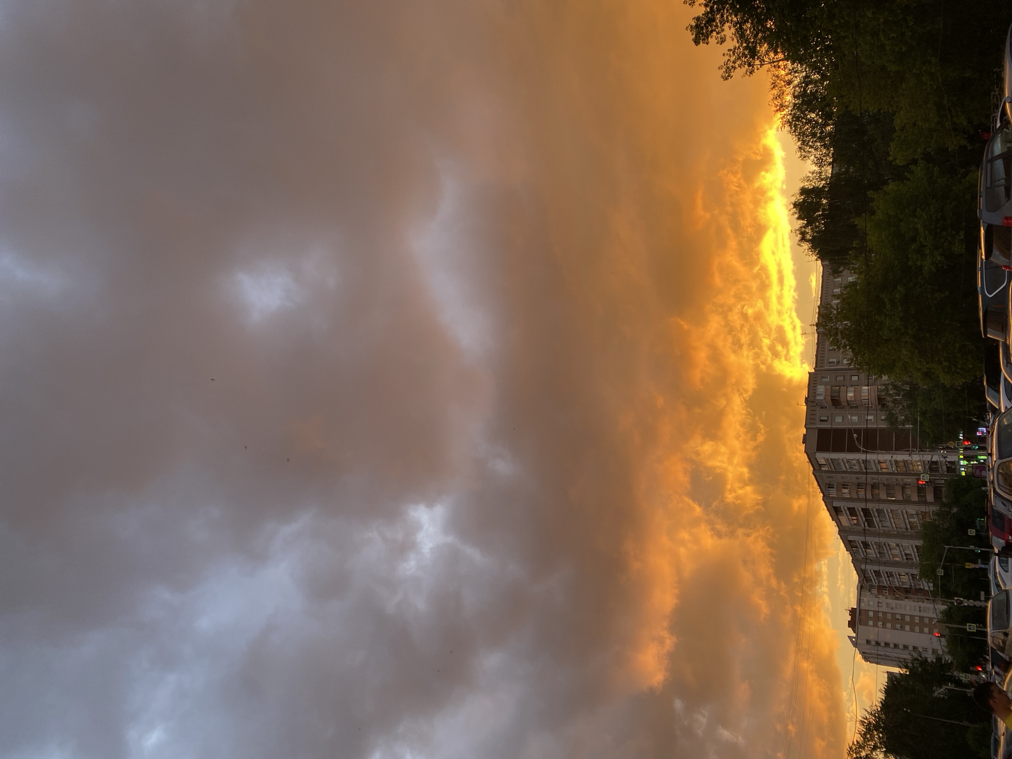 The image, correctly oriented, depicts a dramatic urban landscape under a striking sunset or sunrise sky in Yekaterinburg, Russia. The upper and central portions of the frame are dominated by a visually contrasting sky. On the left, heavy, dark grey clouds create a somber atmosphere, while on the right, the clouds are intensely illuminated in vibrant shades of orange and yellow, suggesting the low angle of the sun. A brilliant, narrow strip of golden light cuts across the horizon line where the sun's rays break through.
Below this dramatic sky, an urban environment is visible. On the left, a line of dark green trees provides a natural silhouette against the bright light. Towards the center and right, several multi-story residential buildings, typical of city architecture, feature numerous windows. Overhead power lines are faintly visible spanning across the sky above these structures. In the foreground on the right, a row of parked cars is partially visible, indicating a street or parking area, with more trees beyond them. A few small, dark specks, possibly birds, are scattered across the cloudy expanse. Traffic lights or street signals with a red light are discernible in the background on the far right. The scene captures a moment of intense natural beauty over a bustling city at either dawn or dusk. No legible text is visible in the image.
