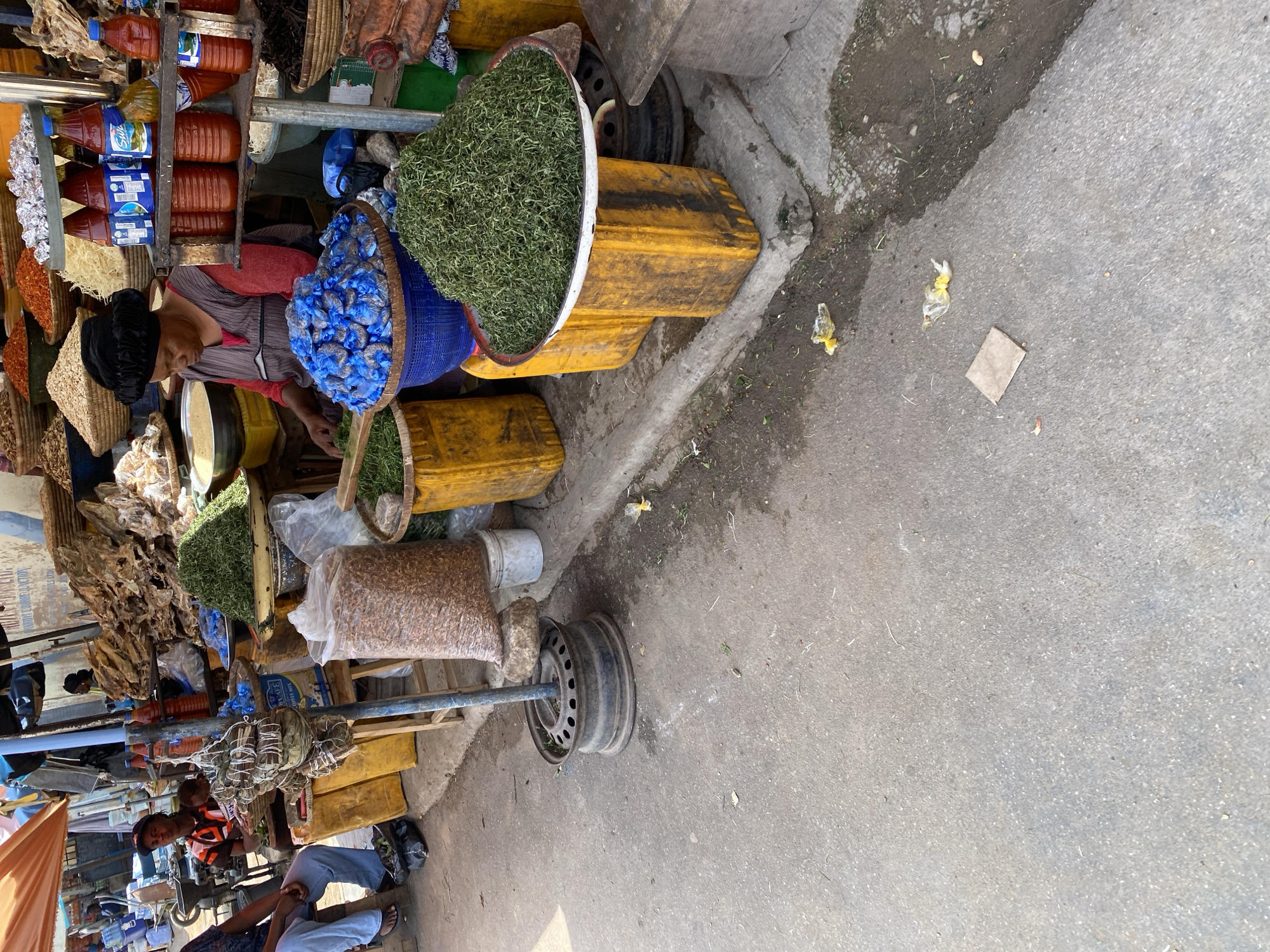 A street market scene in Jos, Nigeria, features a vendor, a woman wearing a black headscarf and a red and brown patterned shirt, tending to her stall. She is situated behind a large blue basket filled with small, blue-wrapped items, and in front of a large pile of dried green herbs displayed on a metal tray. The market environment is characterized by various stalls with goods for sale. To the left of the woman, a person in light blue pants and a dark shirt sits on a stool. In the background, other stalls are visible, displaying an assortment of goods including dried fish, grains, and bottles of sauces or condiments. Yellow plastic containers and sacks of produce are scattered throughout the scene. The ground is paved with asphalt, showing signs of wear and tear, with some scattered debris and a discarded tire rim serving as a base for a market sign. The overall atmosphere suggests a bustling, open-air marketplace.
