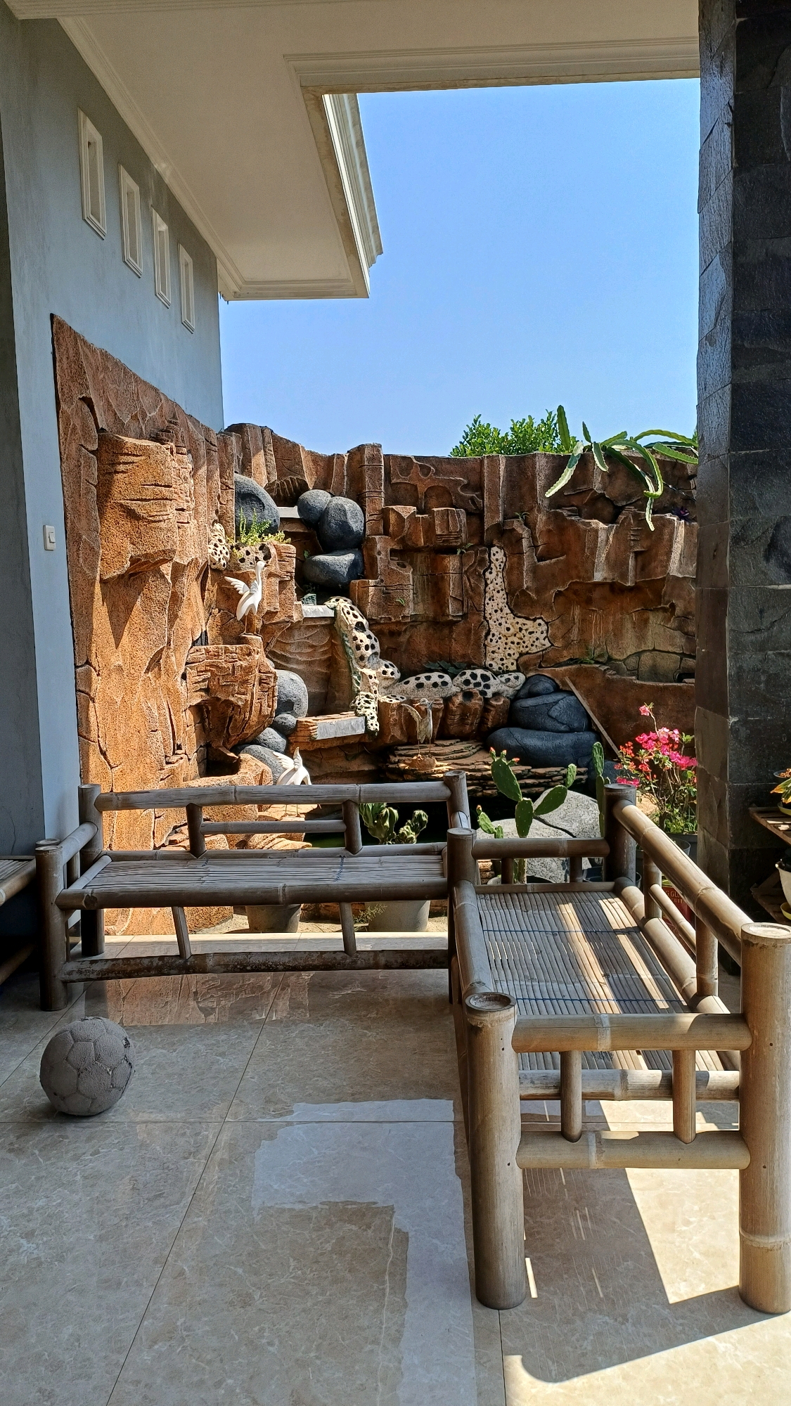 The image depicts an outdoor seating area in front of a decorative rock wall with a faux waterfall and animal figurines. Two bamboo benches are placed on a tiled floor. To the left of the benches, a gray, slightly deflated soccer ball rests on the floor. The rock wall features various textures and colors, resembling a natural cliff face with carved-out sections where the figurines are placed. These figurines appear to be stylized animals, possibly leopards or jaguars, and a white bird. Green foliage and succulents are visible growing from the rock wall and in pots around the area. The scene is illuminated by bright sunlight, casting shadows on the floor and benches. The sky above is clear blue. The architectural elements on the left and right suggest this is an outdoor patio or courtyard area attached to a building. The overall impression is of a decorative and somewhat whimsical outdoor space.