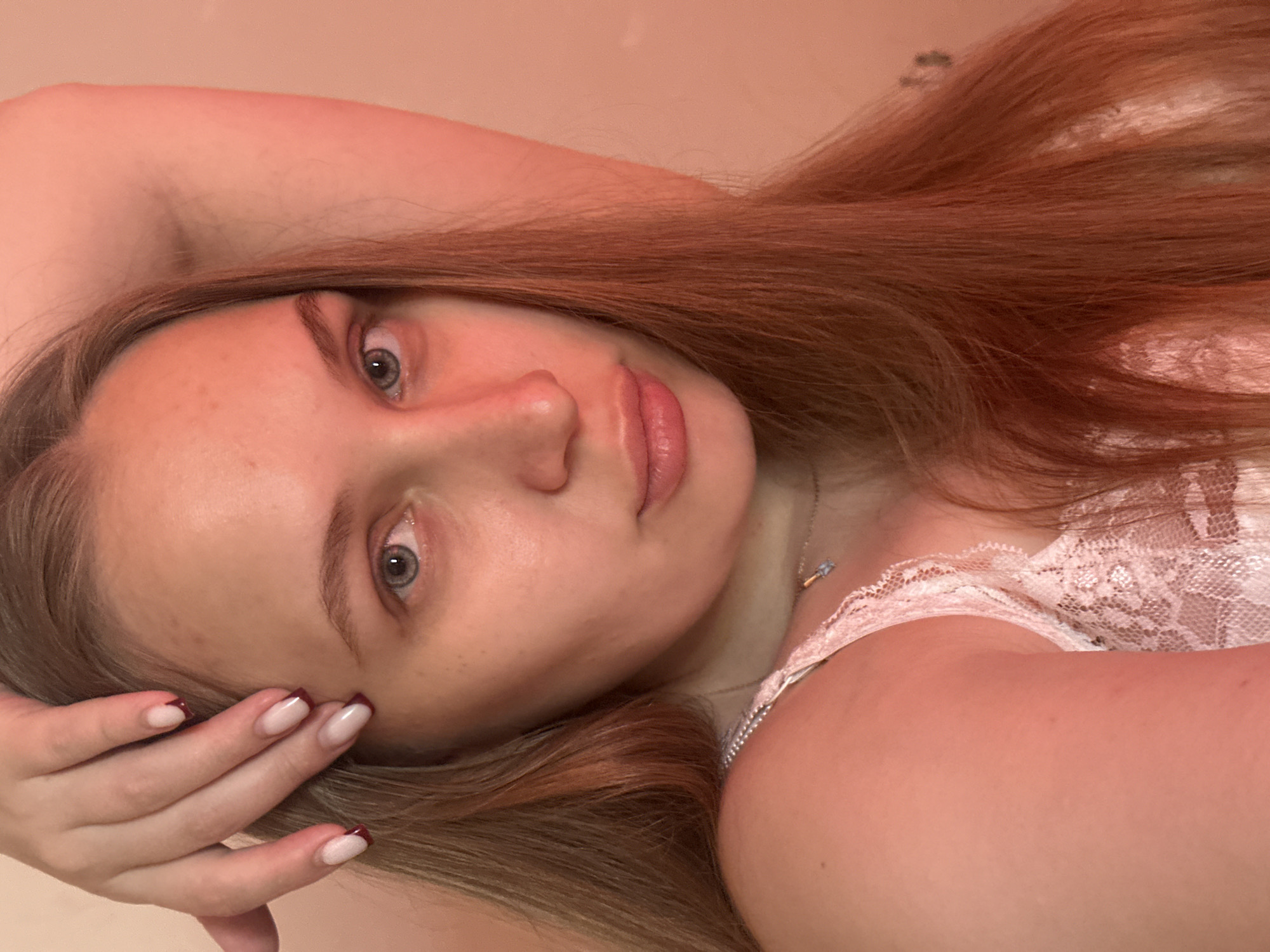 A young woman lies on her side, looking directly at the camera. She has light skin, grey eyes, and auburn hair. Her nails are painted with a French manicure design that includes a maroon tip. She is wearing a light-colored lace top and a thin silver necklace. The background is a solid light pink color, possibly a wall. The location is unspecified, and there is no information to suggest this image was taken in Chaikovsky, Russia.