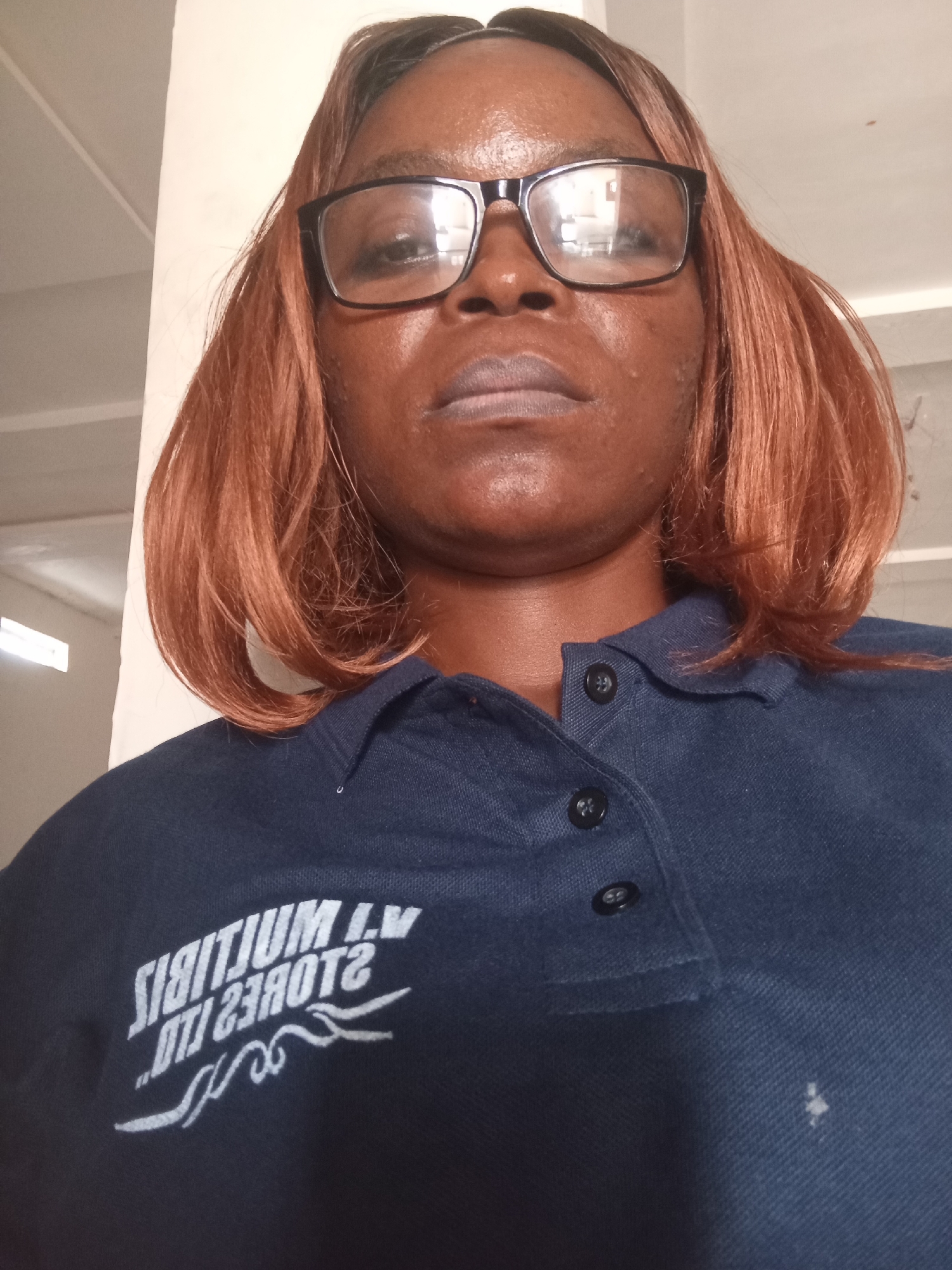 A close-up selfie, taken from a low angle, shows a Black woman wearing glasses and a navy blue polo shirt. She has auburn hair styled in a bob. The shirt has a white logo with text that appears to be backward. The background is a plain wall and a ceiling, with some indistinct structures visible in the distance. There is a faint stain on the shirt, to the right of the logo. The lighting is somewhat harsh, creating reflections on the glasses and highlighting the woman's facial features. The overall setting appears to be indoors.