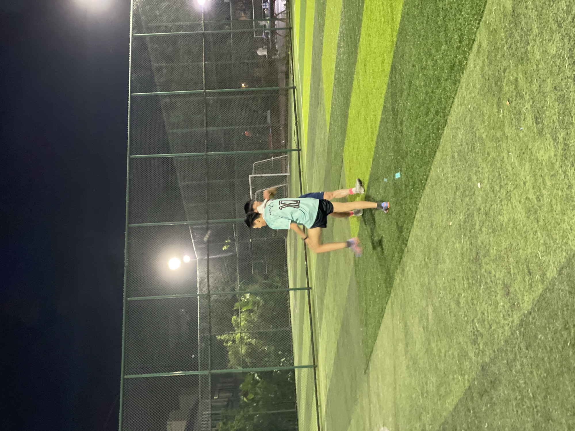 Scene: An outdoor artificial turf soccer field at night.
Location: Likely in the city of Jiangjin, China.
People: Two individuals are present, apparently playing soccer. One wears a light green jersey with a number 11 on the back and black shorts. The second person is partly obscured.
Objects: A soccer goal is visible in the background. The soccer field is striped with varying shades of green. A chain-link fence surrounds the field.
Actions: The two individuals appear to be running.
Environment: It is nighttime, illuminated by artificial lights. There are trees and buildings visible beyond the fence.