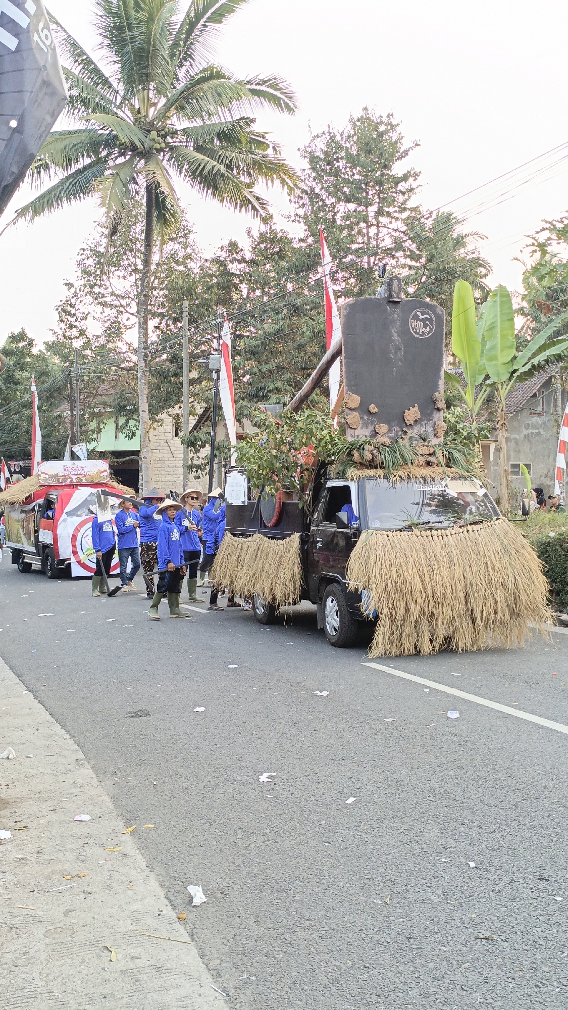 In Krajan Lor, Indonesia, a parade is underway. A black truck, decorated with rice stalks and foliage, drives down an asphalt road. The truck is adorned with a large, dark cylindrical object topped with what appears to be dirt clumps. Several people in blue shirts and hats are walking alongside the truck and another decorated vehicle, a smaller truck with a red and white facade. They are holding farming tools and some are wearing straw hats. Indonesian flags are visible in the background, along with palm trees and residential buildings. The road is littered with debris.