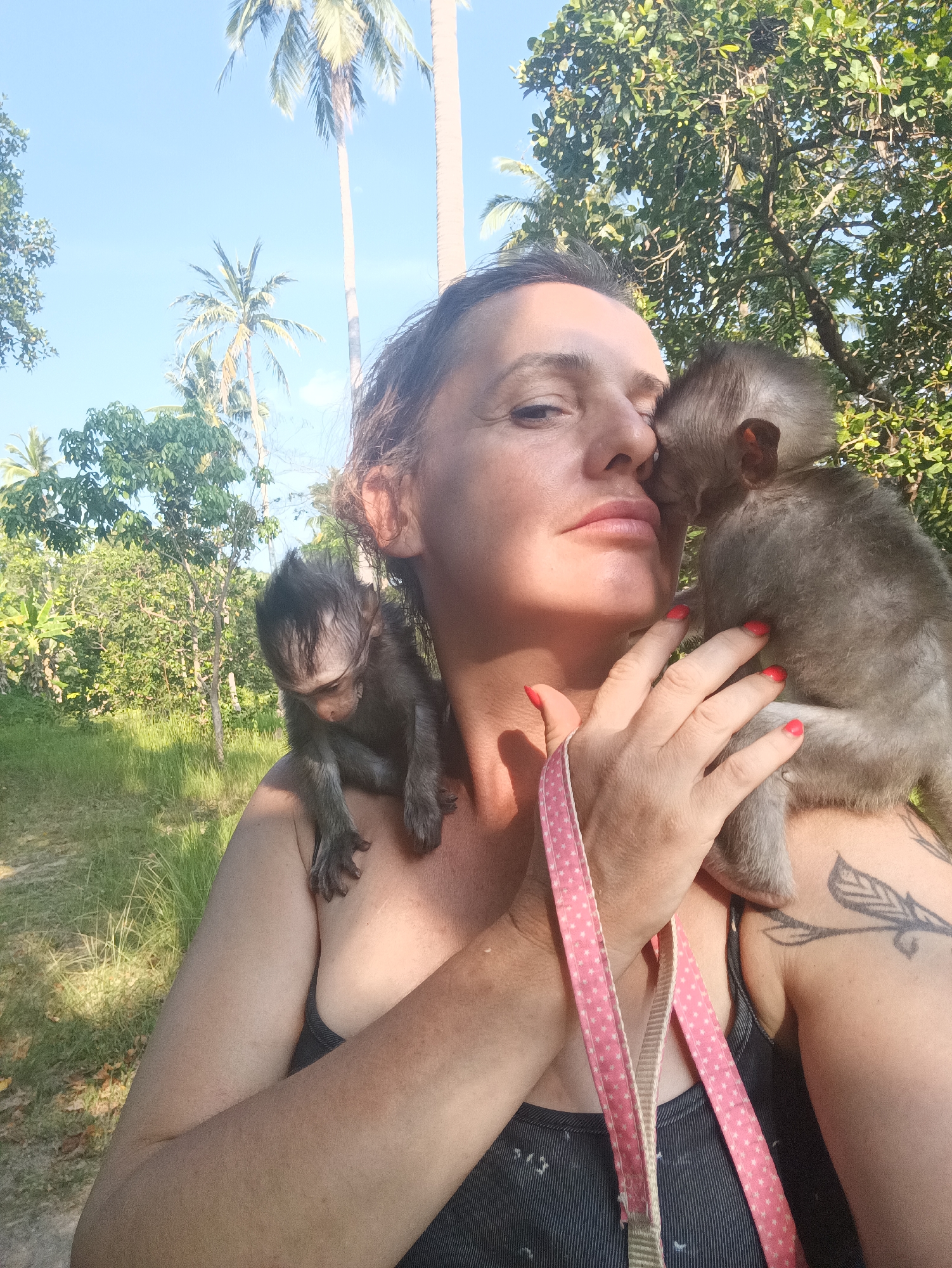 A woman with tan skin, dark hair, and red nail polish is taking a selfie outdoors. She is wearing a dark, sleeveless top and has a pink lanyard with white stars around her neck. A tattoo of a leaf or feather is visible on her right shoulder. Two baby monkeys are perched on her shoulders. The monkey on her right shoulder is nuzzling her face. The monkey on her left shoulder is looking down.
The background includes lush green vegetation and palm trees, indicating a tropical environment. The location is Baan Thong Nang, Thailand, as inferred from the prompt. The time of day appears to be morning or early afternoon based on the lighting. The scene suggests an interaction between a person and wildlife in a natural setting.