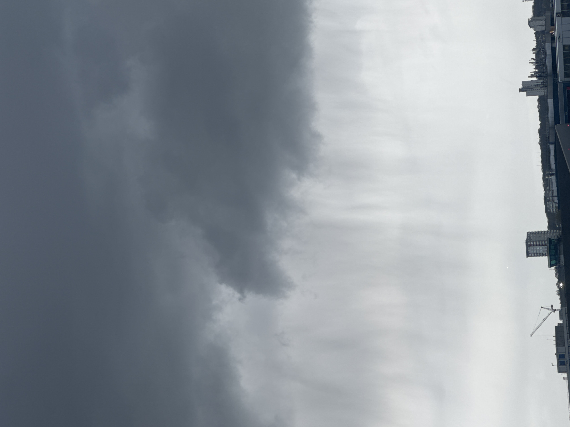 The image shows an overcast sky, with dark storm clouds dominating the left half of the frame. The right half presents a brighter, albeit still cloudy, sky. In the lower right corner, the cityscape of London is visible. Construction cranes and buildings are present, indicating urban development. No people are visible. The environment suggests a potentially inclement weather condition over an urban area.
