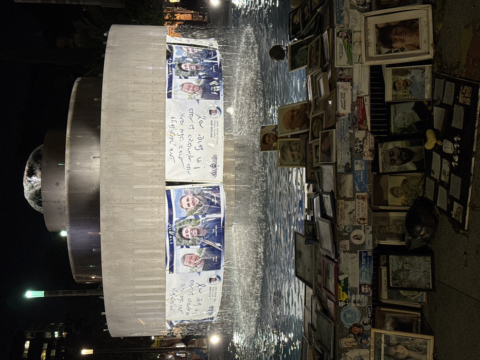 A memorial site at night features a tiered fountain. Posters with images of people and handwritten notes in Hebrew adorn the fountain. Adjacent to the fountain, a collection of framed photographs, stickers, and memorabilia are arranged on the ground. The lighting is dim, with some illumination from streetlights. Some water is visible in the fountain.