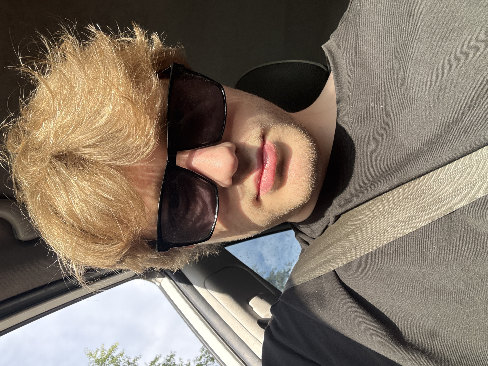 A young person with light blond, messy hair and sunglasses is sitting in the passenger seat of a car. They are wearing a black t-shirt and a beige seatbelt. The sun is shining through the window, casting shadows on their face and shirt. The background shows a cloudy sky with trees visible outside the car window. The location is not specified as being in Krasnogorsk, Russia.