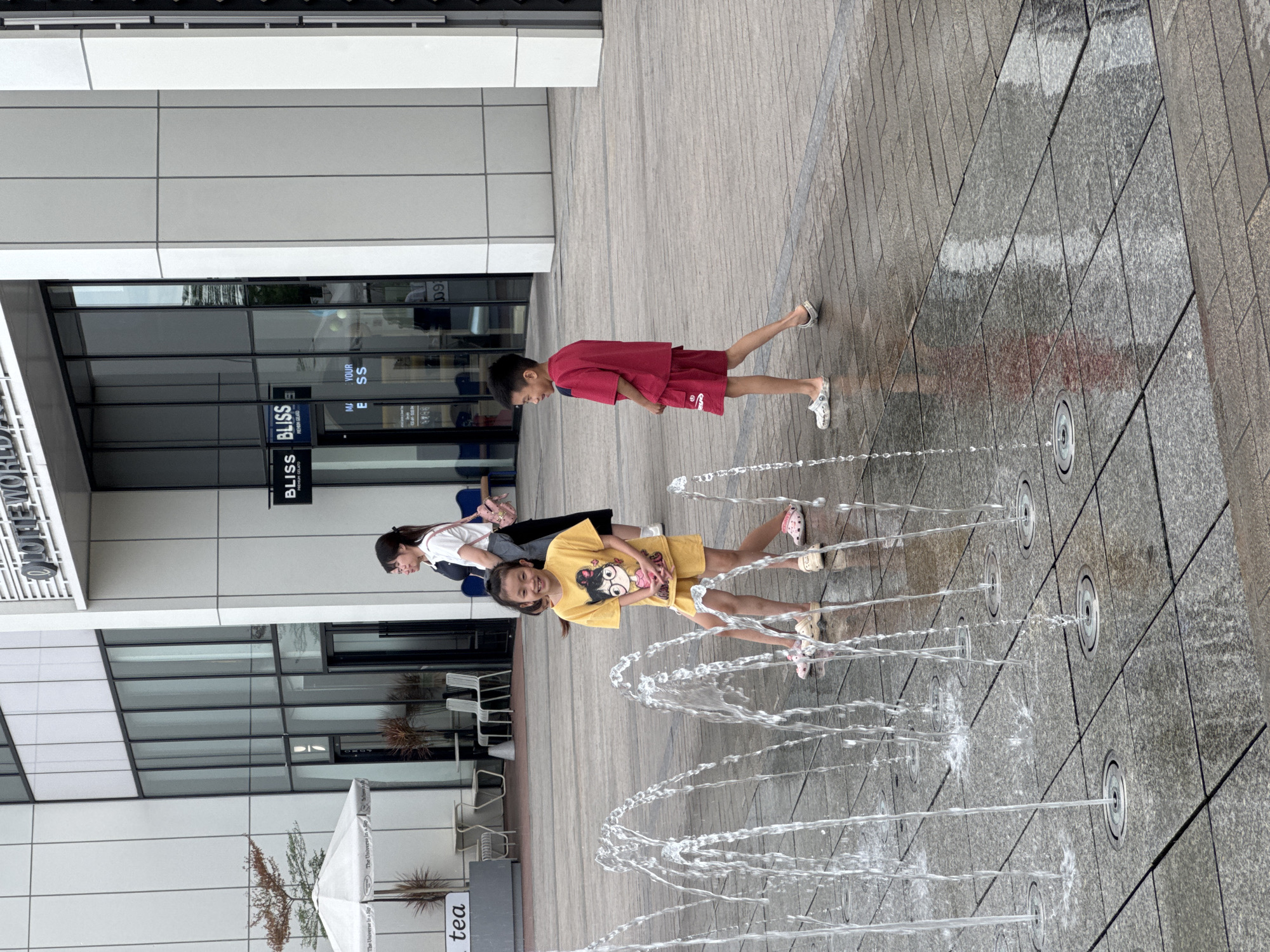 In Hanoi, Vietnam, two children, a girl and a boy, are playing in a water fountain area located in an urban plaza. The girl, wearing a yellow t-shirt and shorts with cartoon prints, smiles as she poses in front of the spraying water jets. The boy, dressed in a red t-shirt and shorts, looks to his left, his hands behind his back. Both children are wearing slip-on shoes. In the background, a woman in a white shirt and dark skirt walks away from the camera, carrying a pink bag. The plaza is paved with grey tiles, and in the far background, a modern building with large glass windows and signage indicating "LOTTE WORLD" and "BLISS" is visible. The area appears to be a public space designed for recreation and leisure, possibly part of a commercial or entertainment complex.