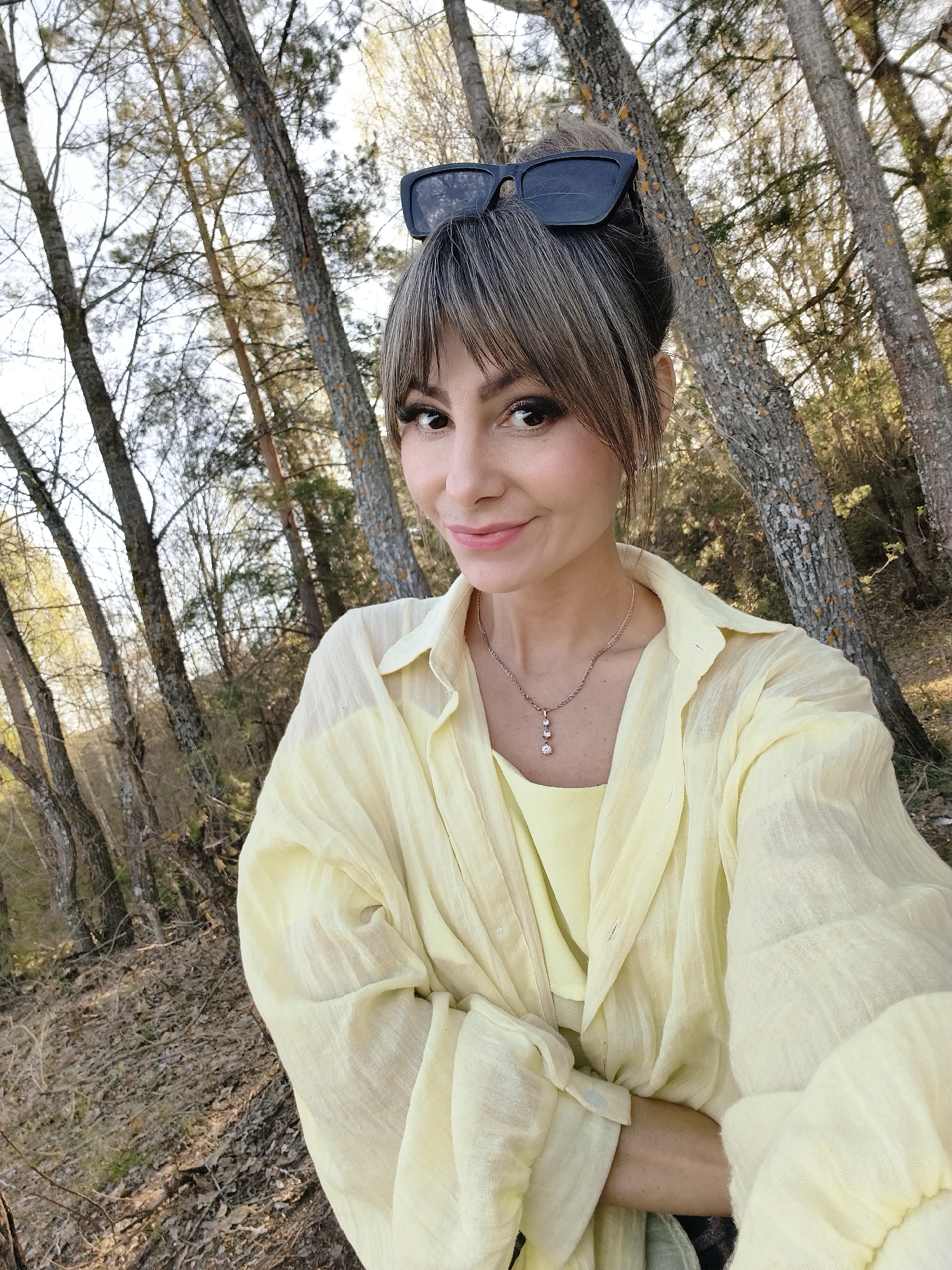 A woman is taking a selfie in a forest. She is wearing a light yellow outfit consisting of a top and a button-down shirt. She has sunglasses on top of her head and a necklace with three pendants. The forest has tall, thin trees. The ground is covered in fallen leaves. The photograph was taken near the city of Chaikovsky, Russia.