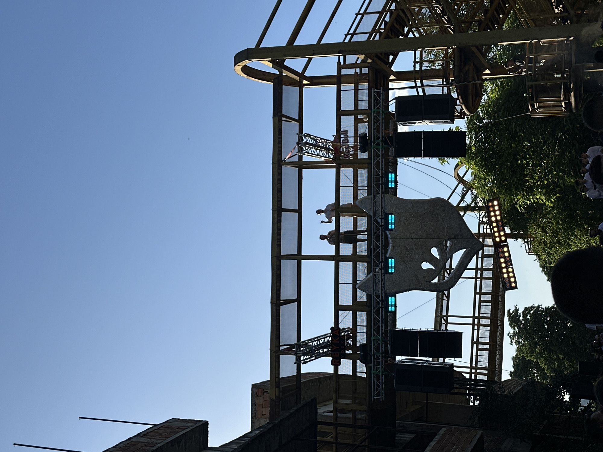 The image captures an outdoor event in Plovdiv, Bulgaria, potentially a concert or festival, set against the backdrop of an industrial-style structure. The structure is made of metal, with scaffolding and mesh panels, and appears to be part of a stage or event installation. A large, artistic sculpture resembling a bear or other animal is mounted on the scaffolding. Speakers and lighting equipment are attached to the structure. Several people are visible on the scaffolding. In the foreground, there are buildings and construction with exposed brick and metal supports. Trees and foliage are also visible, adding to the outdoor setting. The sky is clear and blue.
