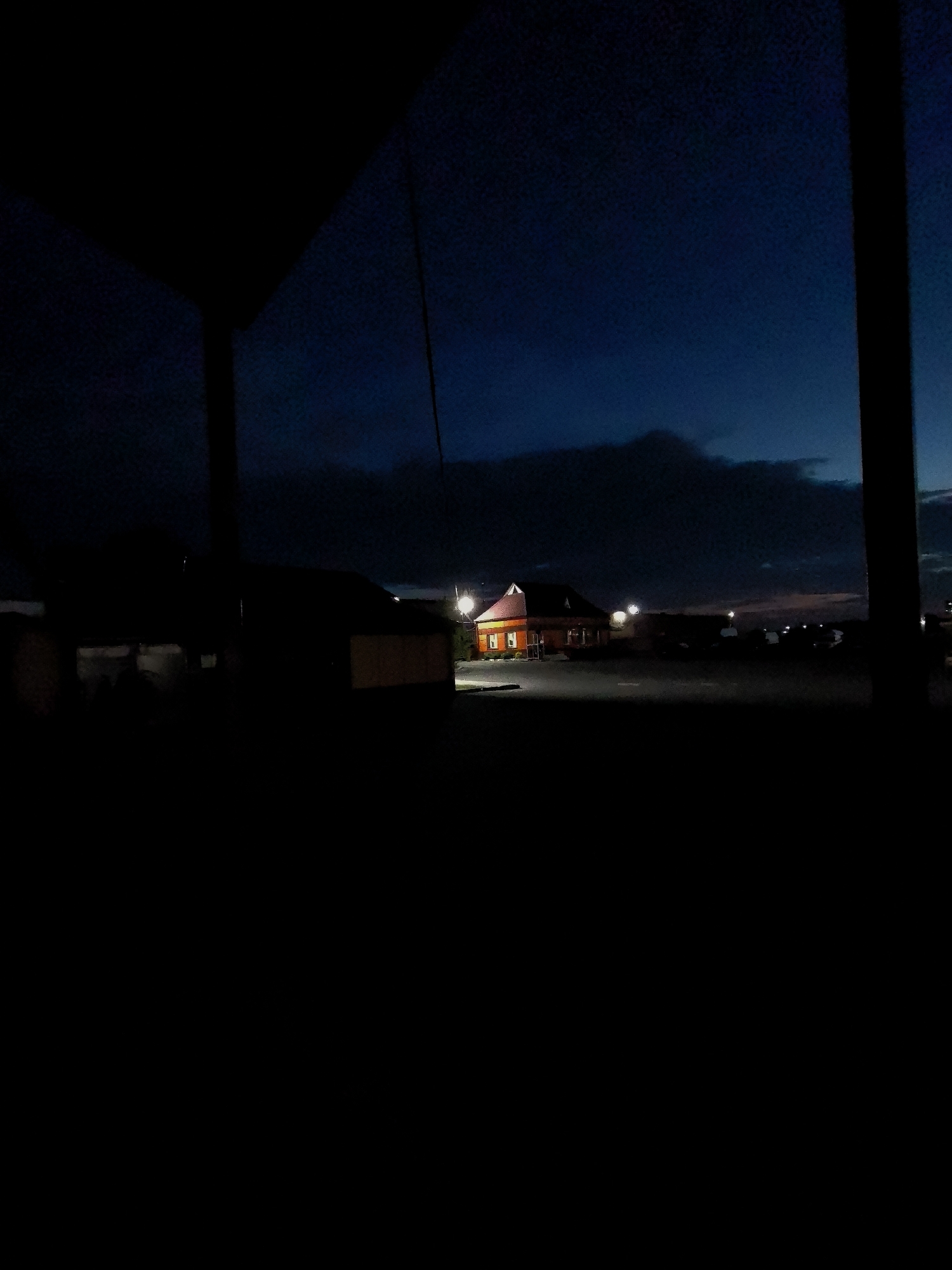 The image depicts a dark, outdoor scene with a building in the distance illuminated by artificial lights. The sky is a deep blue, suggesting it is either dusk or dawn. A dark, silhouetted structure, possibly a roof or overhang, occupies the foreground, obscuring the lower portion of the scene. In the middle ground, a bright orange building with a red roof stands out against the dim surroundings. The building appears to be a commercial establishment, with several windows visible. Exterior lights illuminate the area around the building and a parking lot. The overall impression is one of a quiet, transitional time of day.
