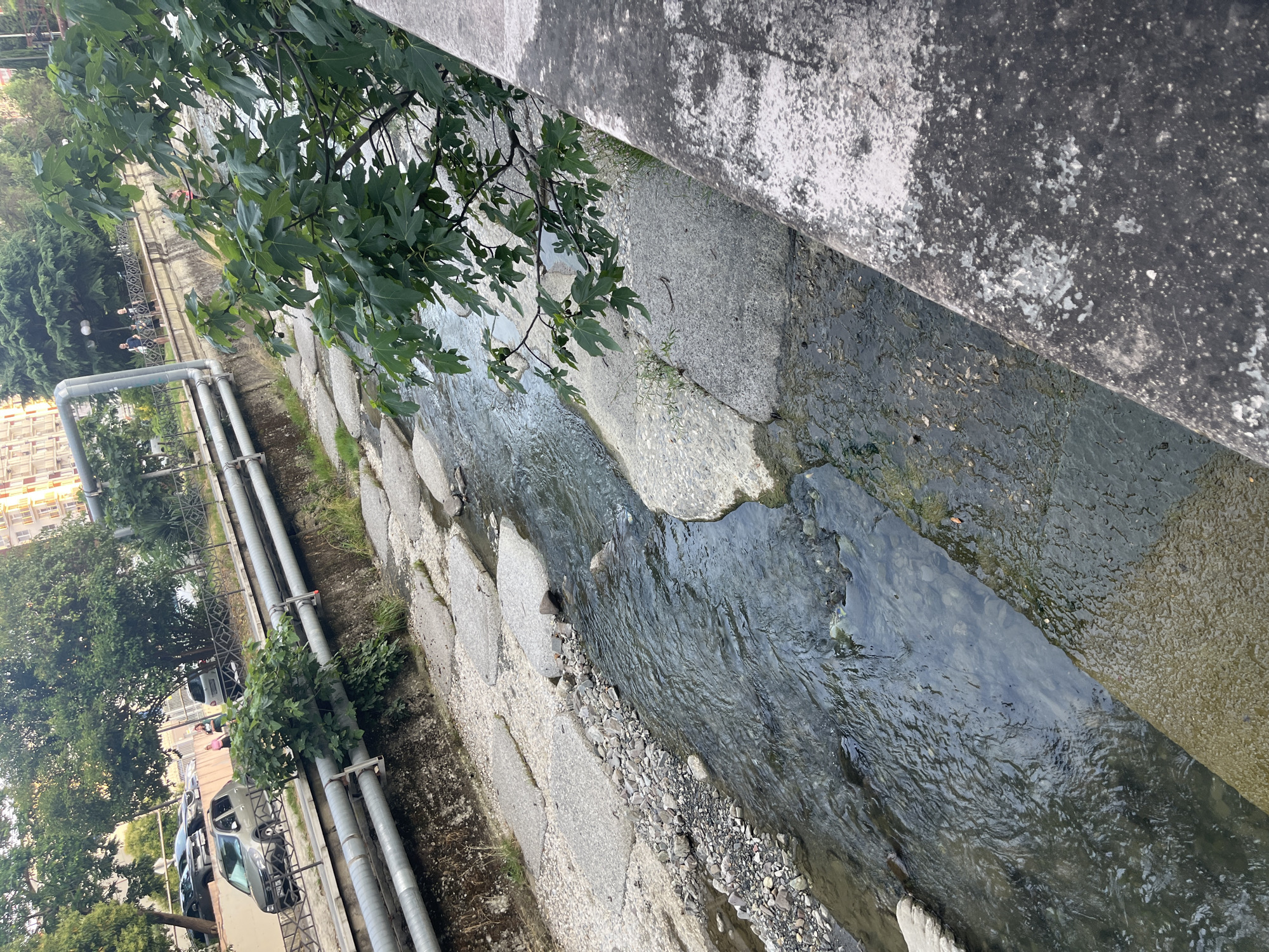 The image depicts a partially submerged concrete canal, with water flowing through it. The canal is lined with large, flat stones and smaller pebbles. Overhanging the canal from the left is a tree with lush green leaves. On the far left, a building with light-colored walls and multiple windows is visible. Adjacent to the building, a row of silver pipes runs along the canal's edge, secured by brackets to a concrete embankment. Further along the embankment, several parked cars and a few people can be seen, suggesting an urban or semi-urban setting. A black metal fence separates the embankment from a pathway or sidewalk where the people are situated. The overall scene appears to be outdoors, possibly during the daytime given the natural light. There is no visible text in the image. The environment suggests a somewhat weathered urban landscape, possibly in Sochi, Russia.