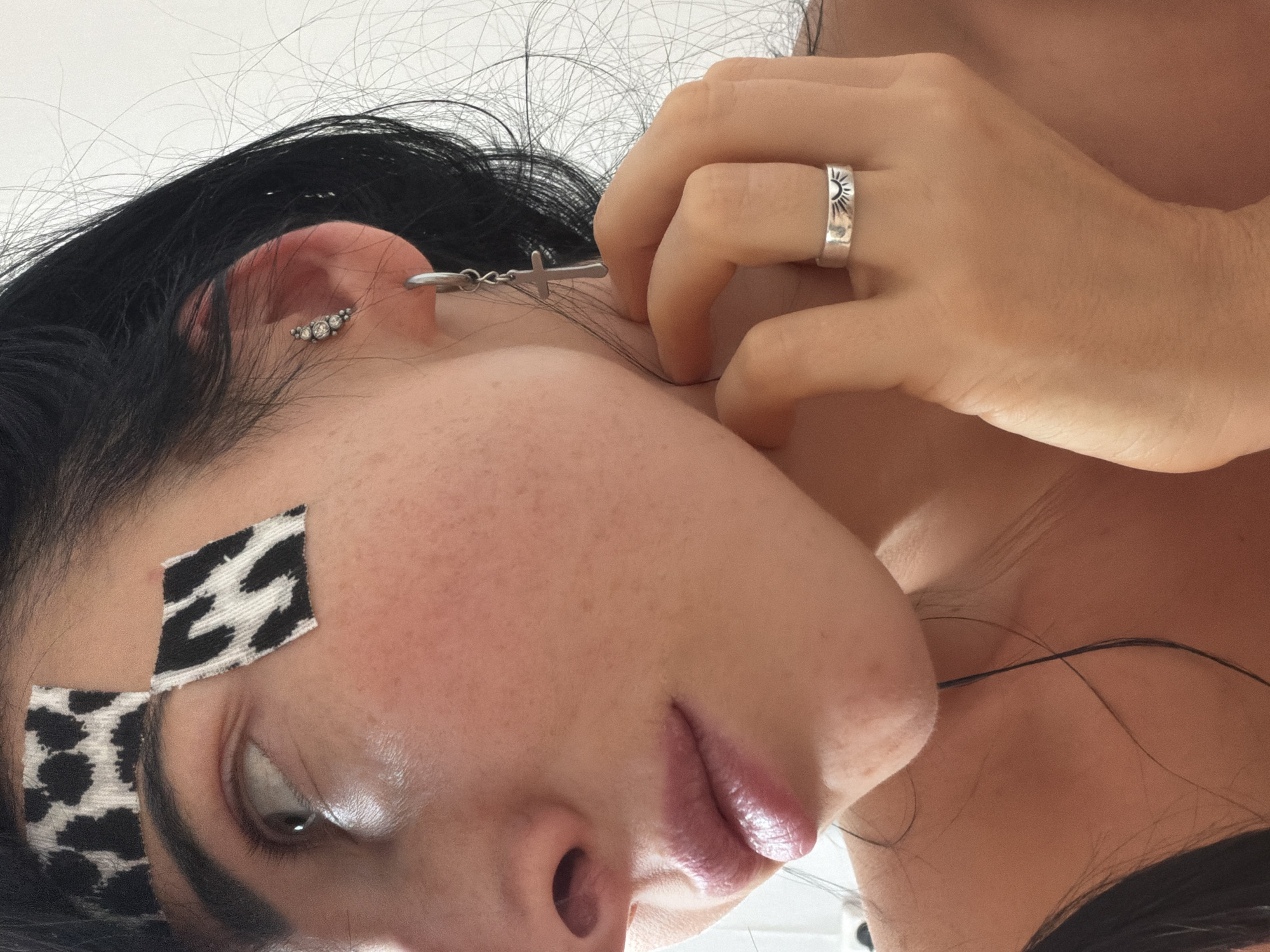 A close-up, high-angle portrait features a person with dark hair and fair skin, depicted from the chest up with their head tilted to the side. The left eye is partially visible, highlighted with a shimmering white eyeshadow in the inner corner. Two distinct diamond-shaped patches, one with a black and white zebra-like pattern and the other with a leopard print, are adhered to the person's forehead/temple area. The lips are slightly parted, showcasing a glossy, pinkish tint.
Adorning the person's left ear are multiple earrings: a curved cluster of small, sparkling stones and a silver hoop from which a small silver cross dangles. The right hand is positioned near the neck/jawline, prominently displaying a silver band ring on the ring finger, featuring an engraved sun motif.
The setting appears to be indoors with a plain white background, likely a wall or ceiling, illuminated by bright, even light. The person's expression is relaxed, with a gaze directed slightly downwards or to the side. The activity primarily involves posing for the photograph. There are no visible location cues within the image itself, nor is any text present. The time of day is indeterminate, though the bright lighting suggests daytime. The context provided indicates the location is Unknown, Russia.