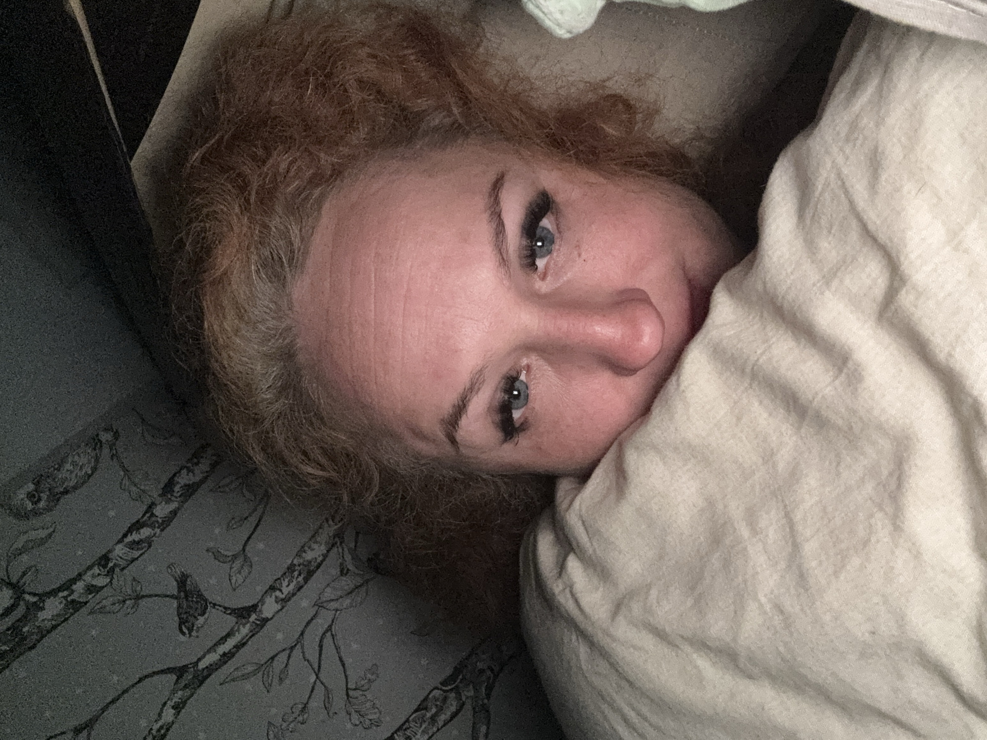 A woman with fair skin and light brown, curly hair is lying in a bed. She is partially covered by a light-colored blanket or sheet. The bedding has a pattern of branches and birds. The location and city of Chaikovsky, Russia cannot be identified from the visual data provided.