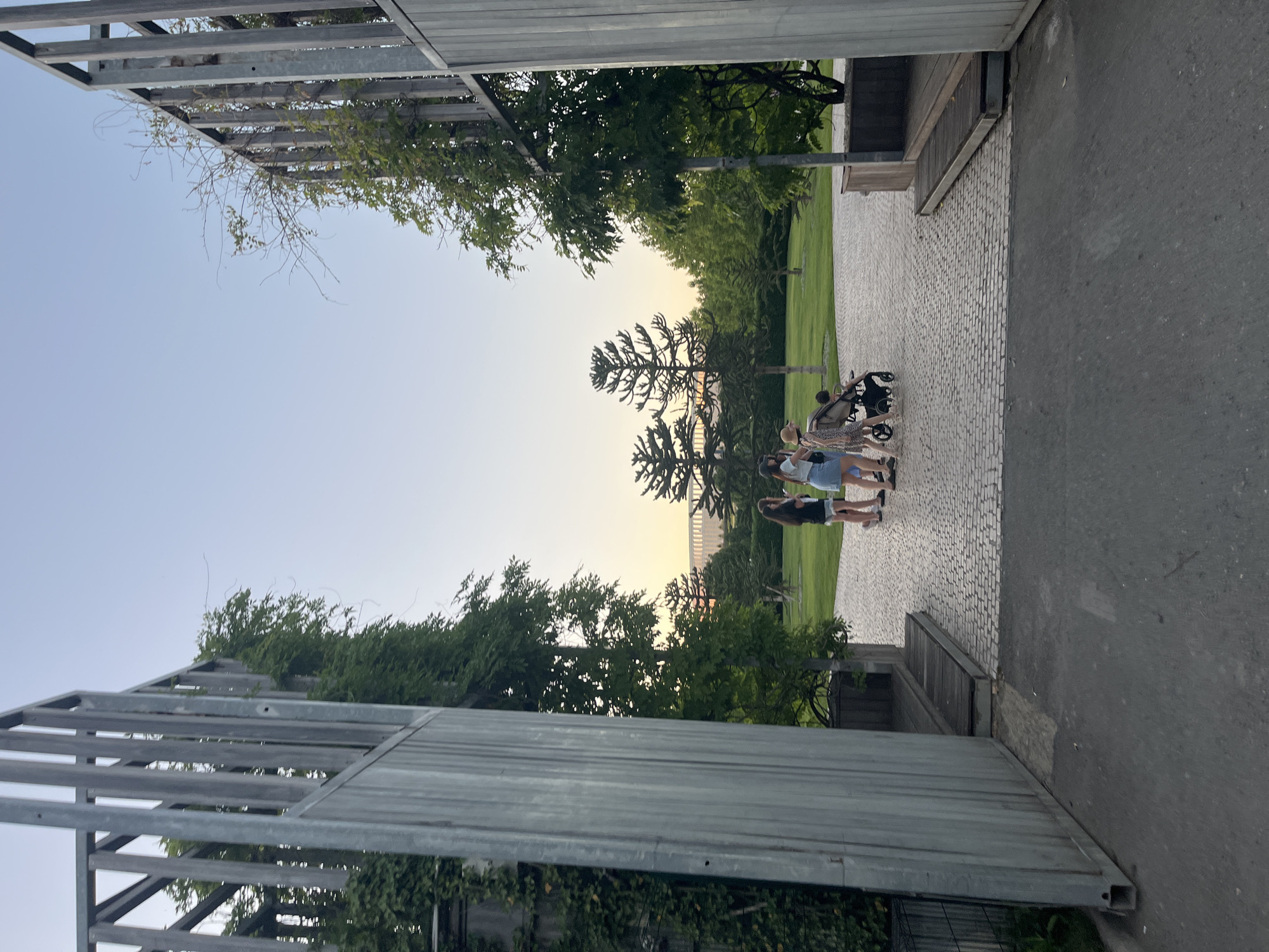 The image depicts an outdoor scene at dusk, likely in a park or public garden, with elements of modern architecture framing the view. In the foreground, two tall, metallic structures with vertical slats stand on either side, creating an entryway or archway. Lush green foliage, possibly ivy, is growing up the left structure. The ground leading into the park is paved with cobblestones.
Further into the scene, a group of people are visible. Four individuals, appearing to be young women or girls, stand together on the cobblestone path. One person is pushing a stroller with a child. The group is facing away from the camera, looking towards the park and the distant treeline.
Beyond the people, a well-maintained green lawn is visible, dotted with trees. In the background, a large, modern building with a curved roofline can be seen, illuminated by a warm glow, suggesting it is lit from within or reflecting the setting sun. The sky above is a clear, pale blue, transitioning to a pale orange near the horizon, indicating sunset. The presence of specific architectural styles, such as the modern structures and the unique tree shapes visible in the park (likely Monkey Puzzle trees), can help identify the location.
This scene is consistent with the atmosphere of the "Krasnodar Park" or "Galitsky Park" in Krasnodar, Russia. The park is known for its contemporary design, extensive landscaping, and striking architectural features, including the iconic illuminated stadium in the background which matches the description. The combination of natural elements and modern art installations makes this a popular public space.