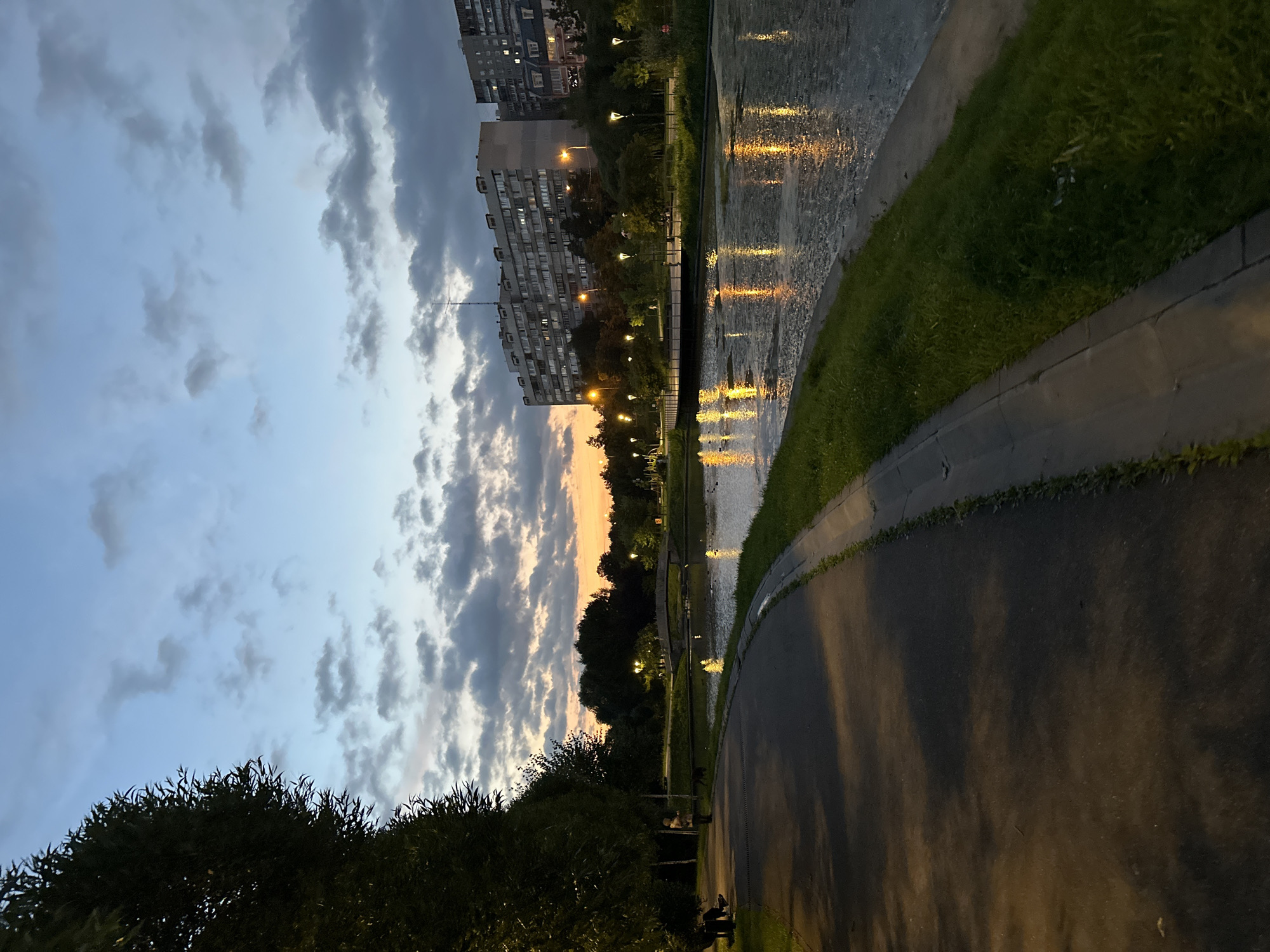 This is a vertical, eye-level shot of a park at dusk. The sky is a mix of soft blue and grey clouds, with a band of orange light on the horizon. In the foreground, a paved path curves to the left, bordered by grass and trees. To the right, a body of water reflects the lights of street lamps and buildings. Several buildings, appearing to be Soviet-era apartment blocks, are visible in the background, illuminated by internal lights. A stone bridge crosses the water in the middle distance, and further beyond, more trees and parkland. There are a few people and a dog in the park, suggesting a public recreational space. The overall atmosphere is serene and twilight-lit. The architecture of the buildings suggests this location is in Moscow, Russia.