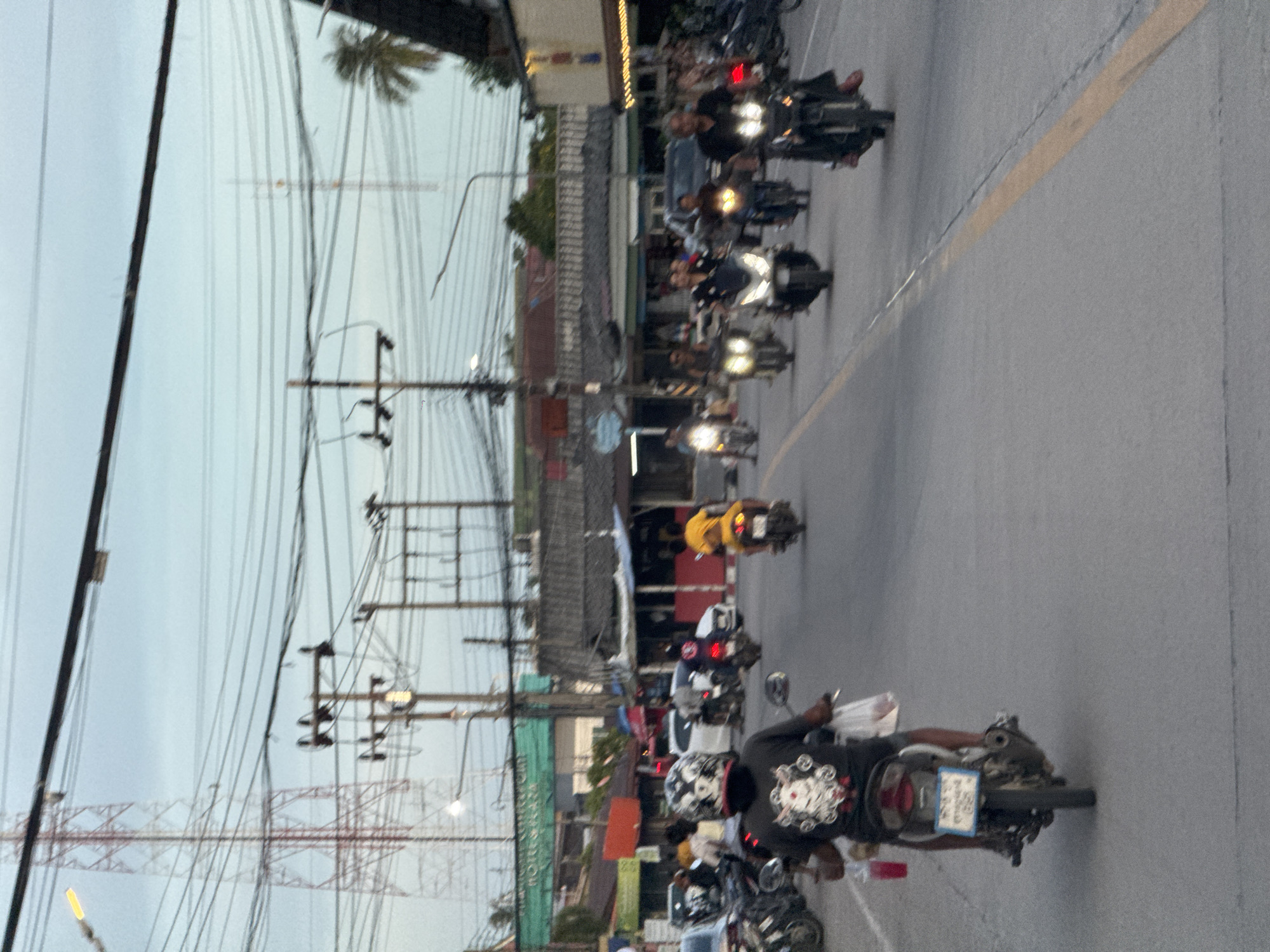 A street scene in Ko Phangan, Thailand, captures a moment of local life with several individuals on motorcycles navigating a paved road. The sky above is overcast, hinting at either dawn or dusk. On the left side of the road, a large red and white electricity pylon stands tall against the sky, with numerous power lines crisscrossing overhead. Adjacent to the pylon, smaller utility poles with insulators are visible. The road is lined with buildings, some appearing to be small shops or residences, with various awnings and roofs. One building on the left has a green sign with white text that is partially obscured.
The foreground is dominated by a person riding a motorcycle, wearing a black t-shirt with a graphic print and a helmet. They are holding a plastic cup with a red liquid in their left hand and what appears to be a plastic bag in their right. Another motorcycle is visible behind them, ridden by someone wearing a yellow shirt. Further down the road, a procession of motorcycles is heading away from the camera, with riders of varying ages and attire. Some are wearing helmets, while others are not. Headlights from some of the motorcycles are illuminated, cutting through the dim light. The overall impression is a typical, bustling street scene in a Thai town, with the omnipresent electrical infrastructure indicating development and connectivity.