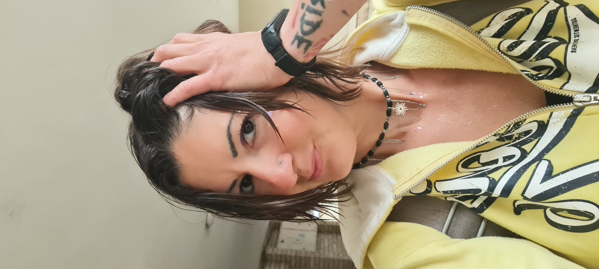 A person with brown hair, a nose ring, and a tattoo on their arm is looking at the camera. They are wearing a yellow zip-up jacket and a necklace. Their hand is on their head. The background is a plain wall. The location is not discernible from the image.