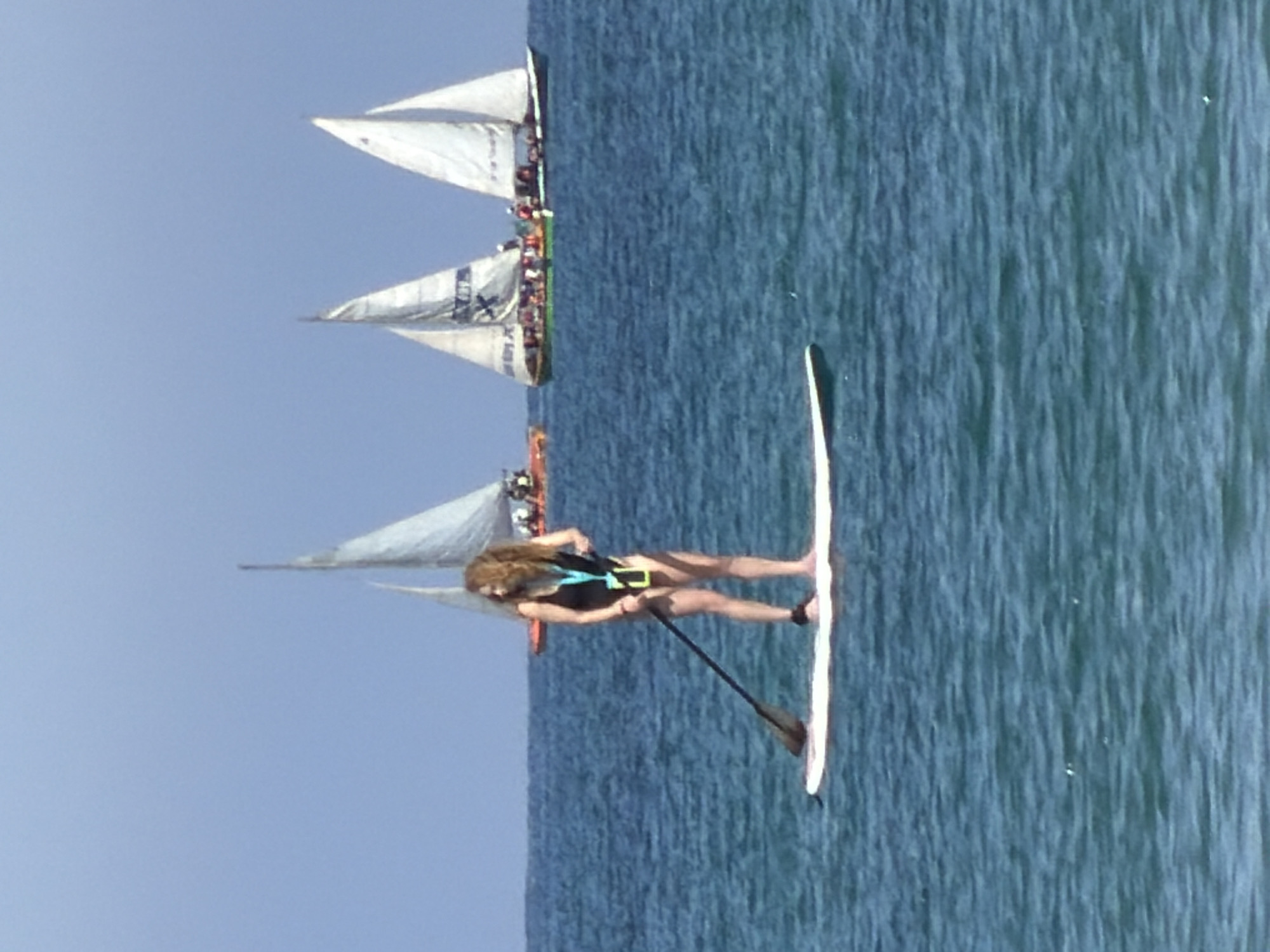 The image shows a woman paddleboarding in the sea off the coast of Tel-Aviv, Israel. She is standing on a white paddleboard and using a paddle to propel herself forward. In the background, there are several sailboats on the water. The sky is blue and clear, and the sea is calm. The sails on the sailboats are white. The water is calm. There is an orange kayak near the woman paddleboarding.