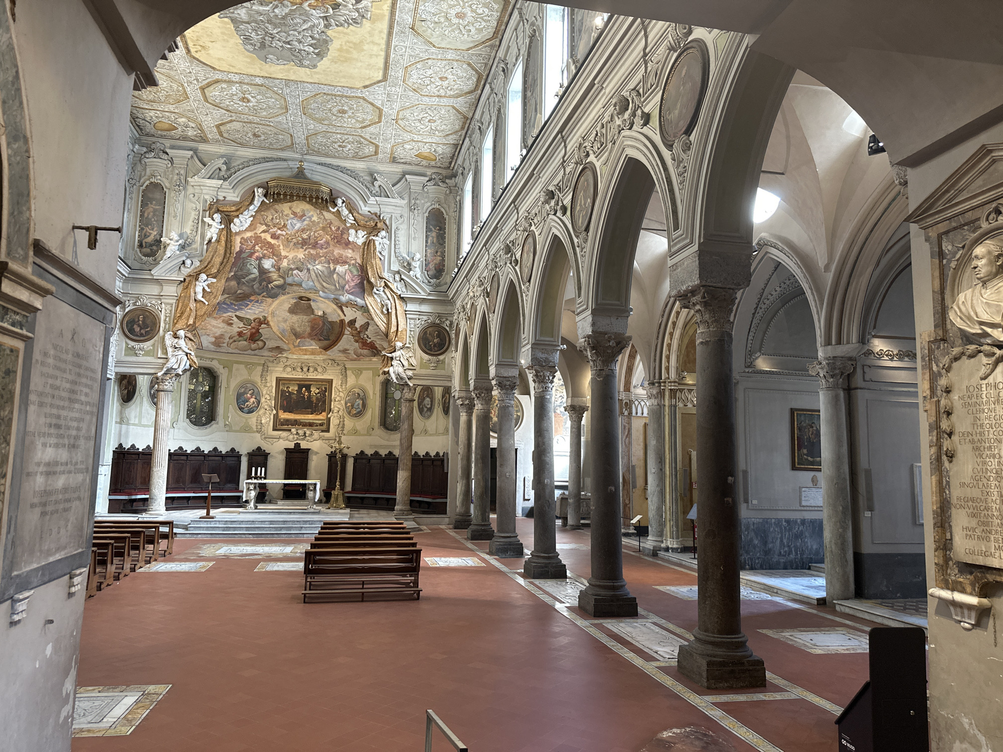 The image shows the interior of a grand church, likely a basilica, with a high, elaborately decorated ceiling featuring frescoes and intricate stucco work. The walls are adorned with paintings and sculptures, and large arched windows allow natural light to filter in. A central nave is lined with tall, fluted columns supporting rounded arches, creating a sense of depth and grandeur. Rows of dark wooden pews are arranged on a red tiled floor, with a raised altar area at the far end. The overall impression is one of opulence and historical significance. This scene is located in Naples, Italy, specifically within the Basilica di San Paolo Maggiore.