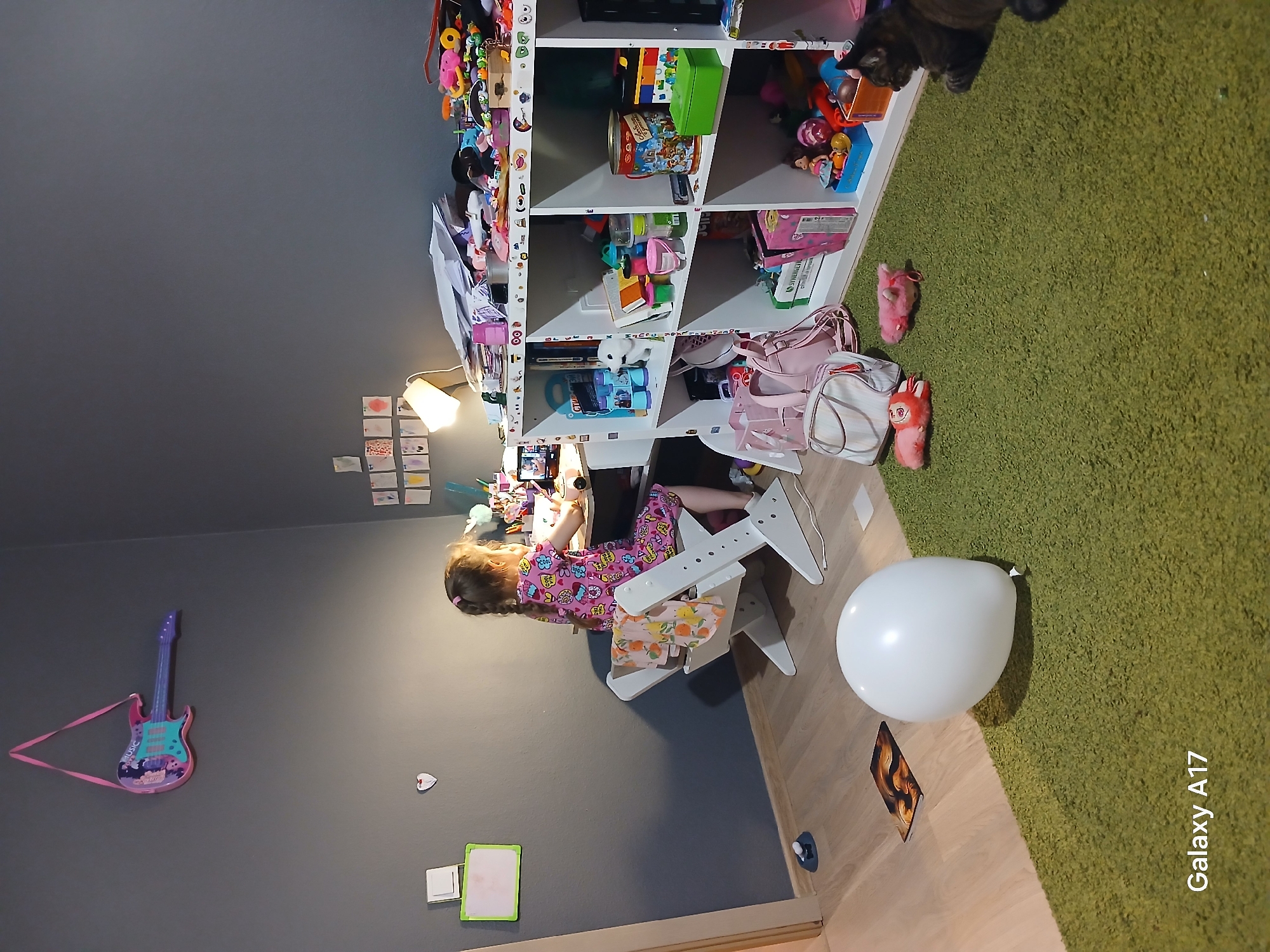 This image captures a child's lively room, likely a bedroom or play area, taken from a slightly elevated perspective looking down and across the space. The room features a dark gray wall on the left and a light wooden floor that transitions into a plush, textured green rug on the right.
The central subject is a young girl, possibly between 4-8 years old, with light brown hair styled in two braids. She is wearing pink pajamas adorned with a colorful, busy pattern. The girl is seated at a white, child-sized desk with a matching white adjustable chair, appearing engaged in an activity with her hands, possibly drawing or playing, under the illumination of a white table lamp.
Behind her is a tall, white multi-shelf unit, overflowing with numerous toys, books, and various colorful containers, reflecting a vibrant play environment. A small tabby cat is partially visible on the top shelf, seemingly resting or observing. Hanging on the gray wall is a pink and purple toy electric guitar with a pink strap. Below it, several small drawings or cards are taped to the wall.
On the floor, near the transition from wood to rug, a large white balloon rests, casting a slight shadow. Further on the green rug, two small pink plush toys are visible, along with a pink handbag and a striped bag. Another small picture or card lies on the wooden floor. The overall impression is one of an active and creative child's space.
A "Galaxy A17" watermark is present in the bottom right corner of the original image (when oriented correctly). The scene appears to be indoors, potentially during the evening or a time when artificial light is needed, as suggested by the illuminated desk lamp and the girl's pajamas. The room's setting is consistent with a home environment in Yekaterinburg, Russia.