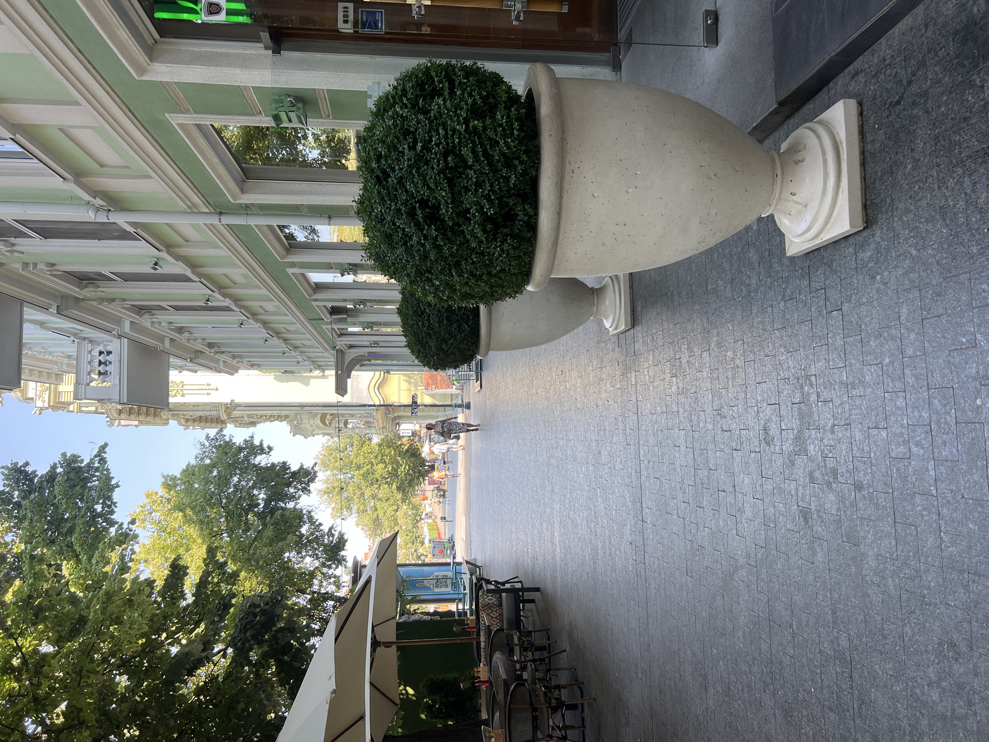 The image depicts a pedestrian walkway in front of a building with light green and white facade ornamentation in Odesa, Ukraine. The walkway is paved with dark grey rectangular stones. On the left side of the walkway, there is an outdoor seating area for a cafe, with tables and chairs visible under a large beige umbrella. Further down the walkway, trees line the left side, and more buildings and trees are visible in the distance.
On the right side of the walkway, two large, light-colored urns are placed, each containing a neatly trimmed, dark green spherical bush. Behind the urns is the entrance to a building, with a glass door and a green neon sign. A woman in a patterned dress is walking away from the viewer down the center of the walkway. The scene is illuminated by natural sunlight, casting shadows on the pavement. The overall impression is one of an urban environment, possibly a commercial or residential street with outdoor amenities.