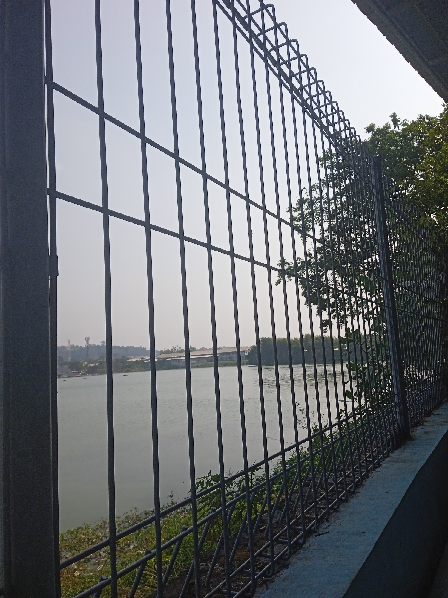 The image shows a view of the Banjir Kanal Barat (West Flood Canal) in Semarang, Indonesia, through a metal fence. The canal is calm and reflects the overcast sky. In the distance, across the canal, are buildings and vegetation, with some telecommunication towers visible. The foreground is dominated by the dark metal bars of the fence and a concrete ledge. The fence has a decorative, wavy pattern along the top. The overall scene suggests an urban waterfront area, possibly a public space or a protected facility along the canal. There are no people clearly visible in the image. The atmosphere is subdued, with muted colors due to the lighting conditions.