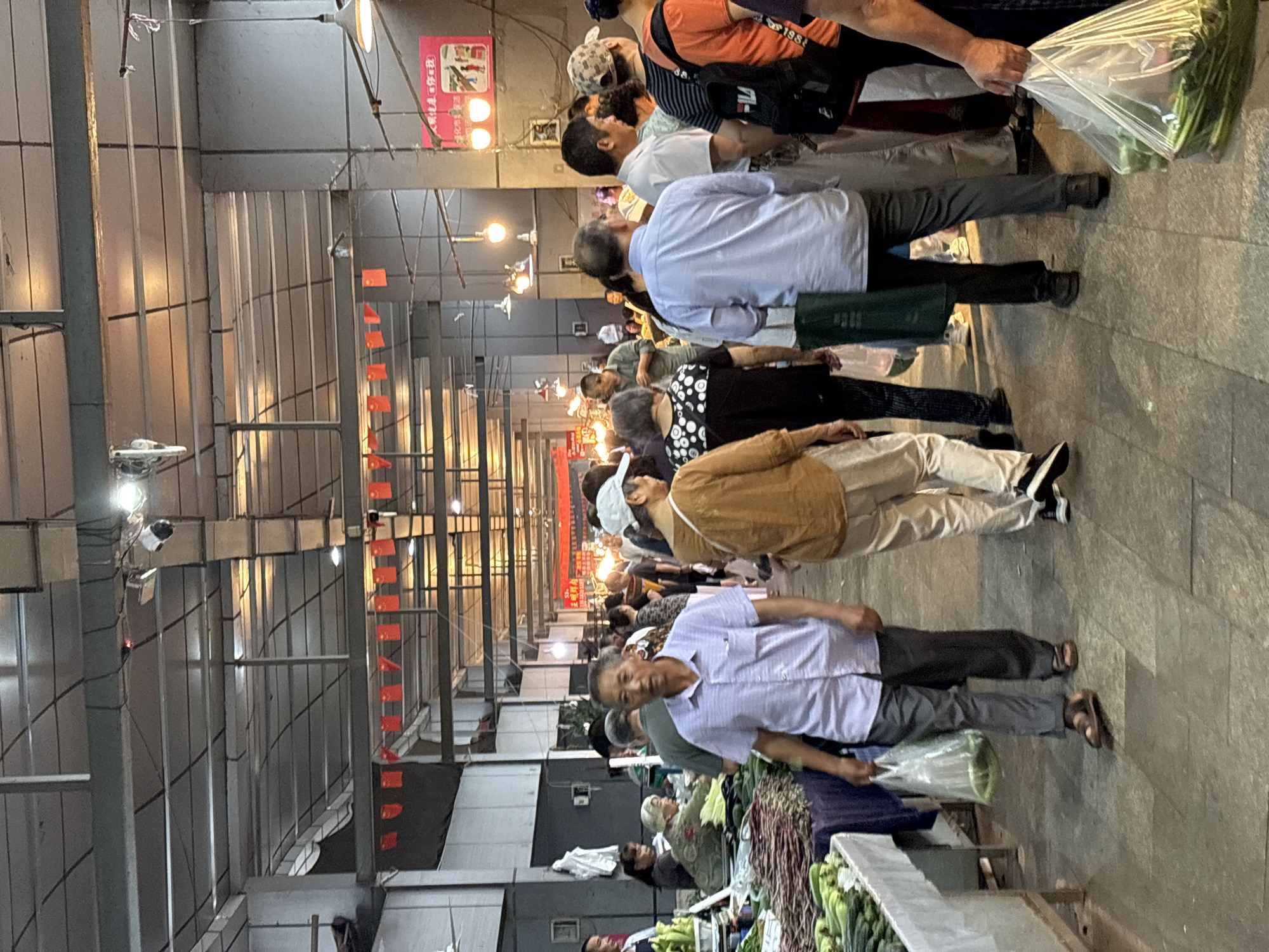 The image shows a crowded market scene, likely in Jiangjin, China. People are shopping at stalls piled with vegetables. The market appears to be covered with a metal roof structure and hanging red flags. The scene suggests a typical day at a local market.