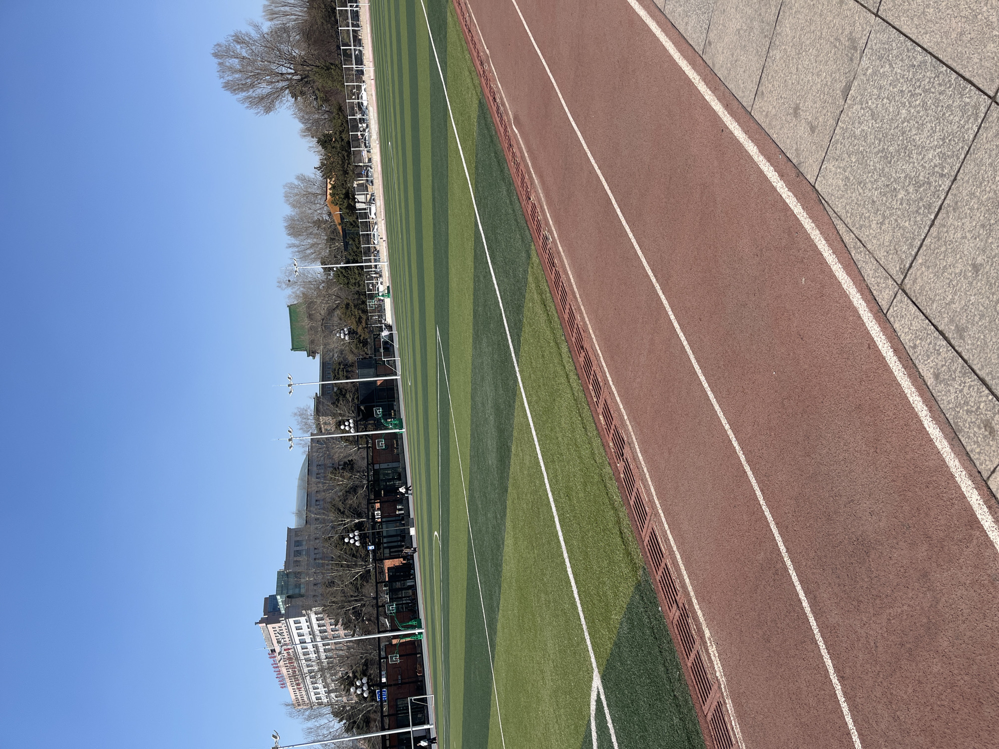 The image presents a view of a sports field and running track in Changchun City, China. The scene is captured under a clear, bright blue sky.
The field itself is green, marked with lighter green stripes, and is flanked by buildings, trees, and streetlights. A red running track borders one side of the field. White lines demarcate the individual lanes. A concrete walkway runs along the edge of the track.
The surrounding architecture appears to be a mix of modern and older buildings. Trees, still barren from the winter, line the street. Streetlights with spherical lamps are visible. The buildings extend along the border of the field.