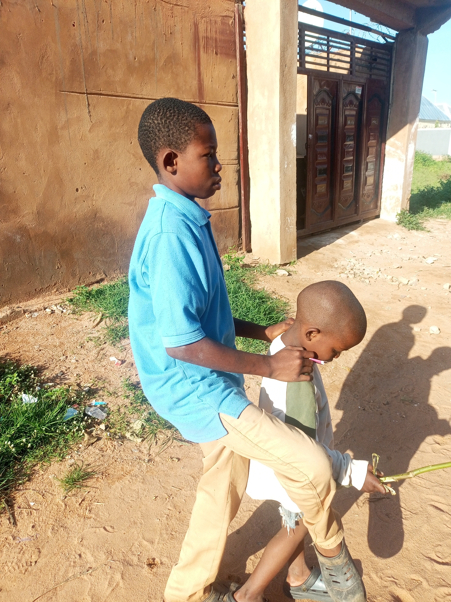 The image captures two young boys in an outdoor setting, likely in the city of Gombe, Nigeria, as indicated by the presence of a rustic, earth-toned building with a large, decorative metal gate, and the dry, sandy ground with scattered vegetation, typical of the region.
The boy in the foreground, facing left and slightly away from the camera, is wearing a light blue polo shirt and tan pants. His stance suggests he might be leaning or assisting the other boy. His hands are positioned around the younger boy's neck and upper back.
The second boy, positioned in front and slightly to the right, is facing down and to the left, seemingly engaged with something in his hands. He is wearing a white shirt with a green stripe and dark-colored Croc-style sandals. His left hand appears to be holding a thin, green stick or stalk.
The environment is characterized by rough, sandy soil, patches of dry grass and weeds, and what appears to be debris on the ground. The sunlight casts distinct shadows of the boys and the surrounding structures onto the ground, indicating a bright, sunny day. The building in the background, with its rough texture and imposing gate, suggests a residential or commercial property with a traditional architectural style. The sky above is clear and blue.