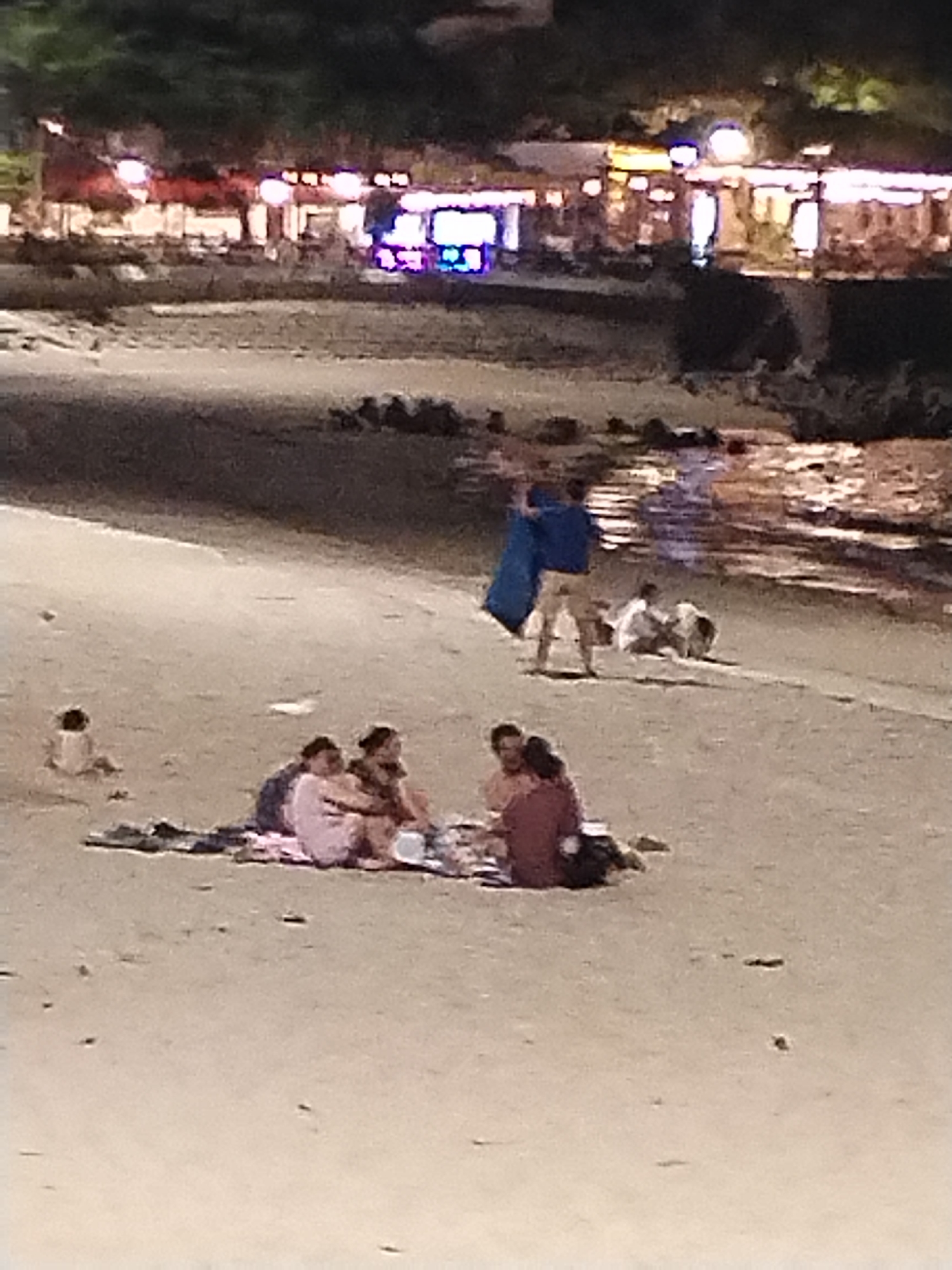 The scene depicts a beach at night in Nha Trang Ward, Vietnam. Four people are sitting on a blanket spread on the sand in the foreground, appearing to be having a picnic or gathering. Behind them, a person is standing and holding a blue object, possibly a surfboard or a large bag. Further back, two other people are sitting or kneeling on the sand near the water's edge. The background shows a blurry view of buildings and lights along the coast. The water reflects the lights, indicating it is a calm evening.