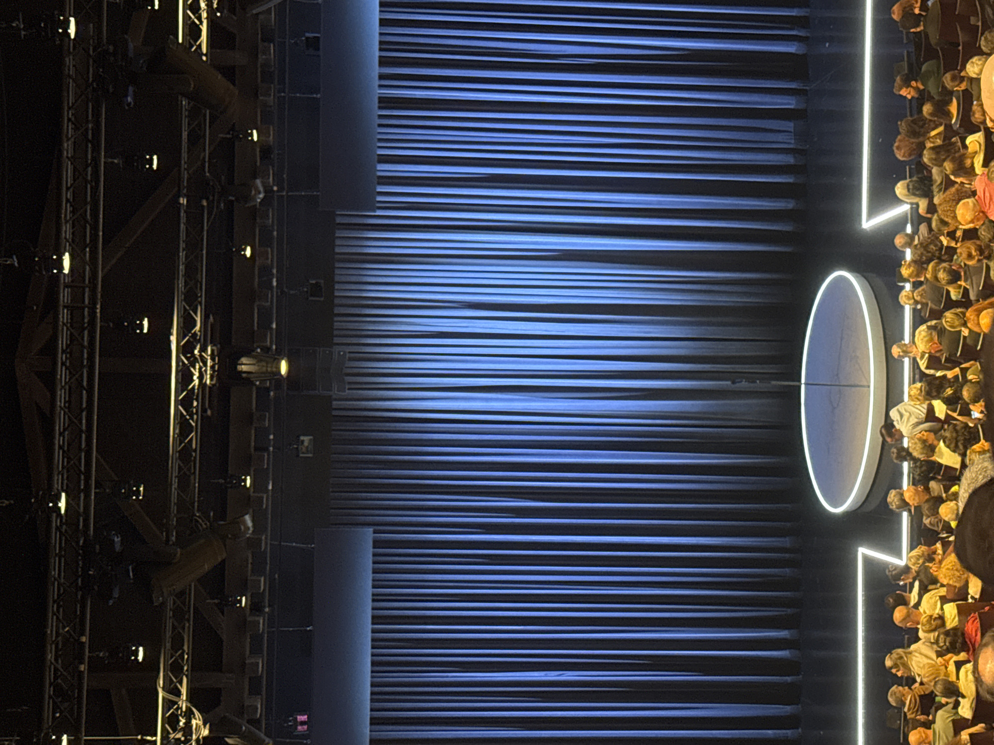 The image depicts a performance stage with a blue horizontal striped backdrop. A circular structure with a white outline is situated on the right side of the stage, near an audience of people seated in rows. Stage lighting equipment is visible on the left. The location is potentially a performance venue in Tel-Aviv, Israel. The presence of an audience suggests a live performance or event.