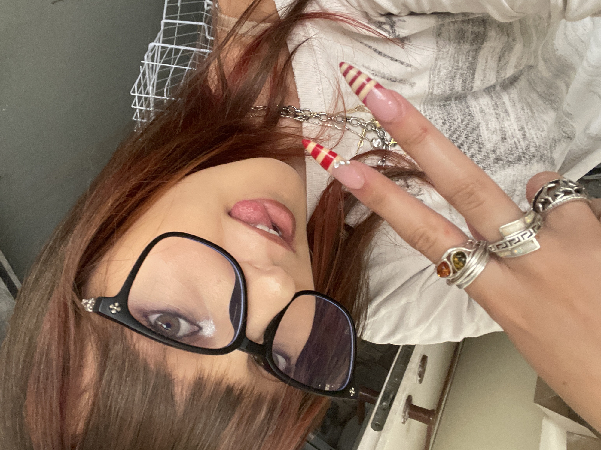 This is a close-up, angled, outdoor selfie of a person in Raanana, Israel. The subject is a young woman with reddish-brown hair, wearing black-rimmed glasses with decorative embellishments on the temples. She is wearing a white t-shirt with gray horizontal stripes. She is making a peace sign with her right hand, which has long, stiletto-shaped nails painted with red and beige stripes and some rhinestones. She is also wearing several rings on her fingers, including one with multiple amber-colored stones and another ornate silver ring. A silver necklace with various charms is visible around her neck. Her tongue is sticking out slightly, and she appears to be smiling. The background is blurred, but appears to be an outdoor setting with some furniture and a metal grate visible.