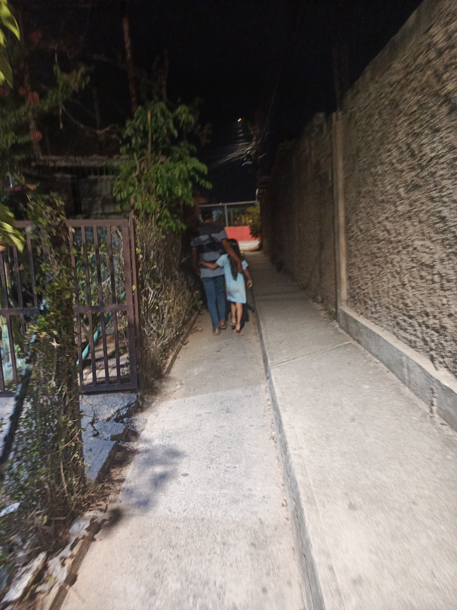 A poorly lit alleyway in Juan Griego, Venezuela, shows a man and a child walking away from the viewer. The man is wearing a striped shirt and jeans, and has his arm around the child, who is wearing a light-colored dress. They are walking down a narrow concrete path between a wall on the right and a metal fence overgrown with vegetation on the left. The alleyway appears to be dimly lit, with the darkness increasing in the distance.
