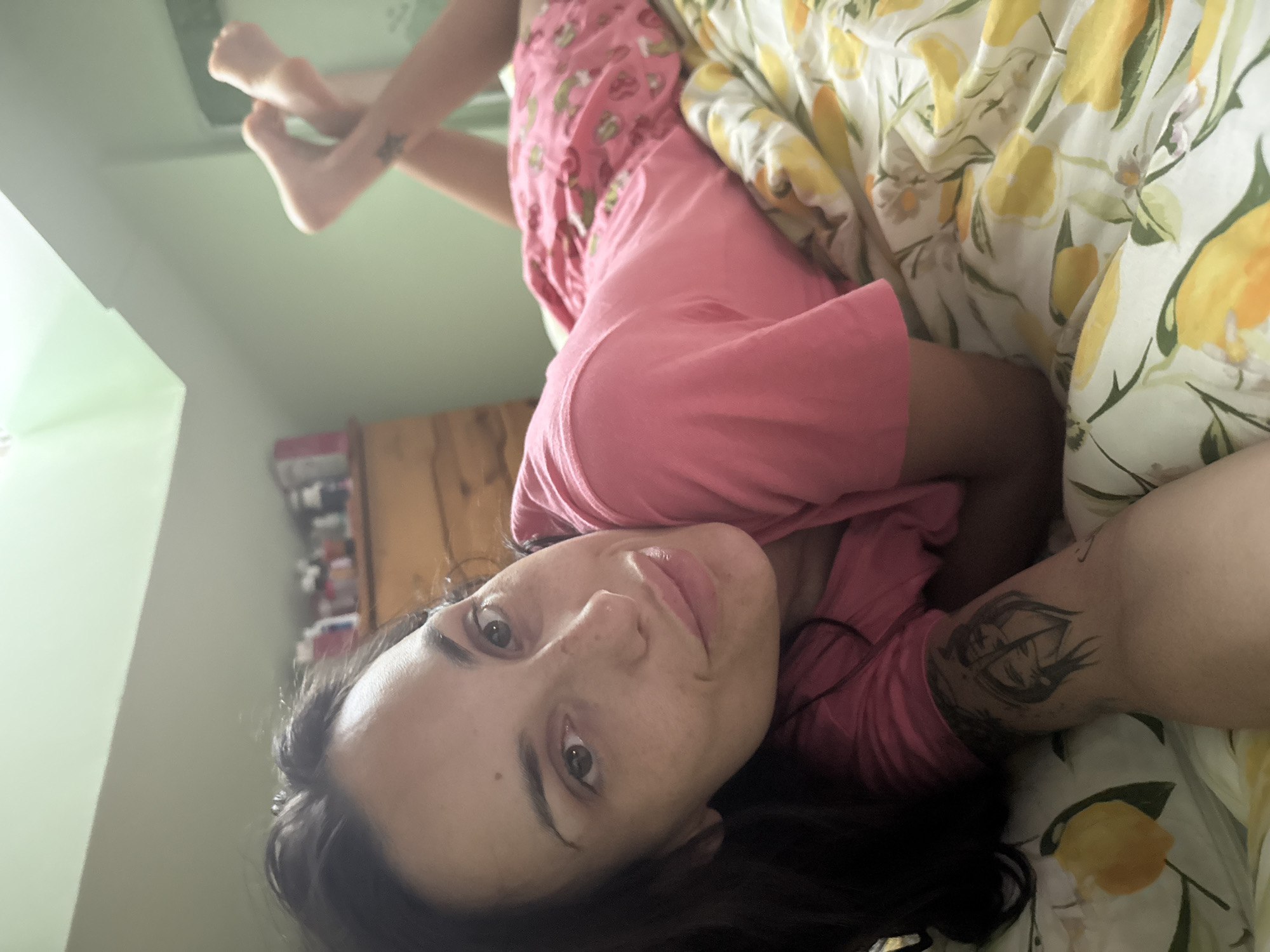 A woman is lying on a bed in what appears to be a bedroom. The woman is wearing pink pajamas. She is looking at the camera. The bed is covered in a white blanket with a yellow lemon print. The room has green walls and a brown cabinet. There is no indication of the location being in the city of Pleven, Bulgaria.