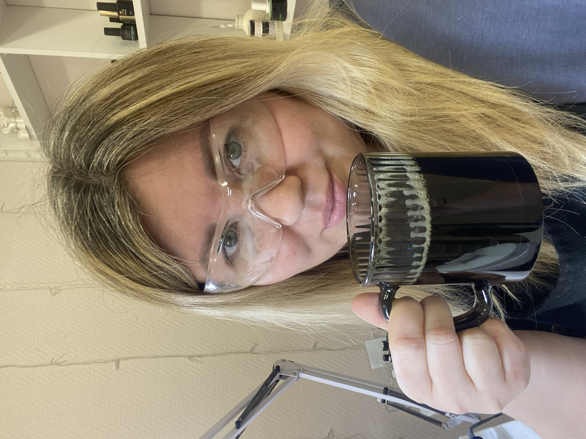 A woman with blonde hair and wearing clear safety glasses is holding a black mug. The background includes a shelf with bottles, a textured wall, and an adjustable lamp. There is no information in the image to indicate a location in the city of Chaikovsky, Russia.