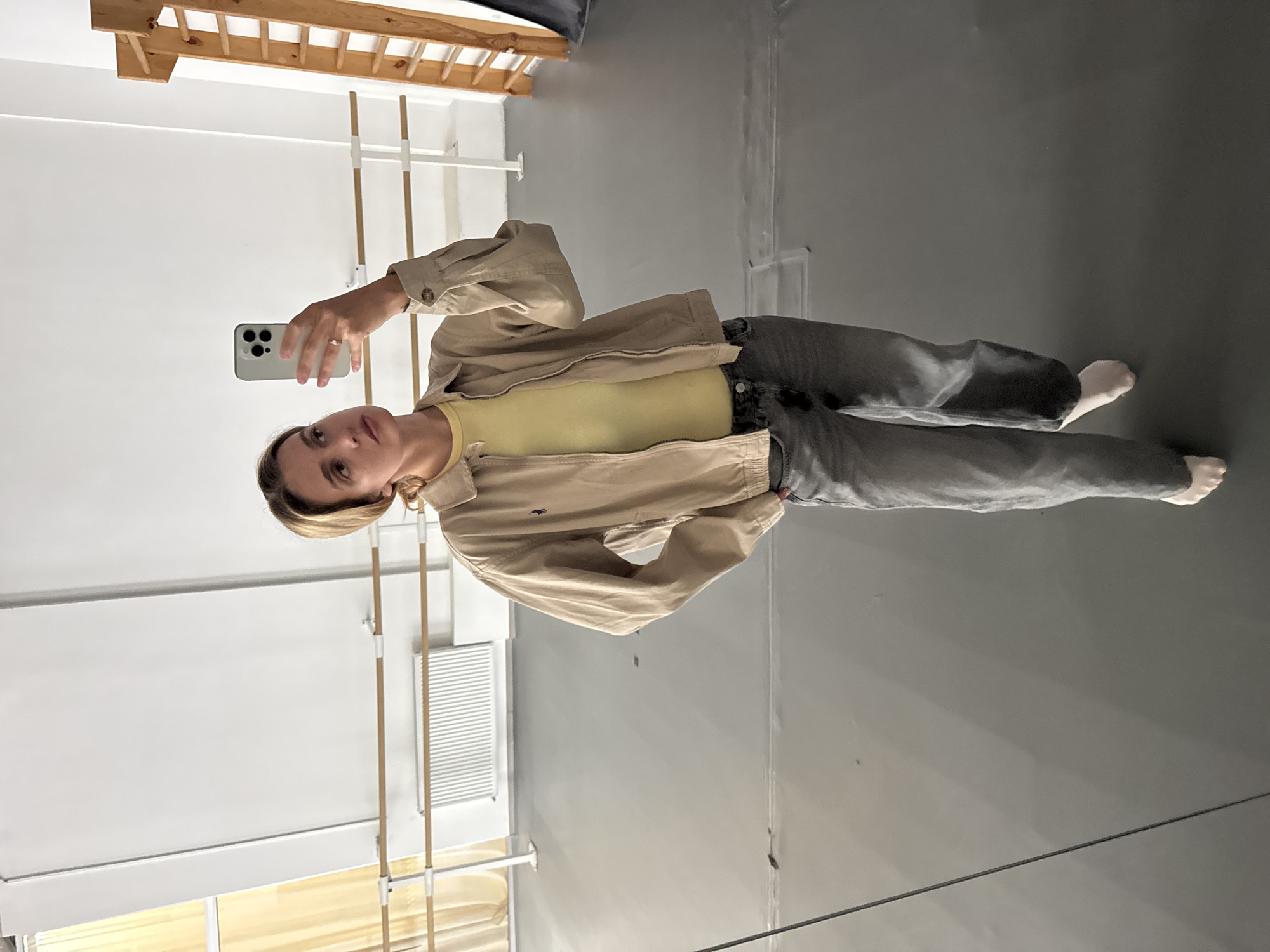 A young woman is standing in a dance studio, taking a selfie in the mirror. She has blonde hair and is wearing a light yellow top with a tan jacket and grey jeans. She is barefoot. The studio has white walls, a grey floor, ballet barres, and a window with a yellow curtain. The location is Unknown, Unknown.