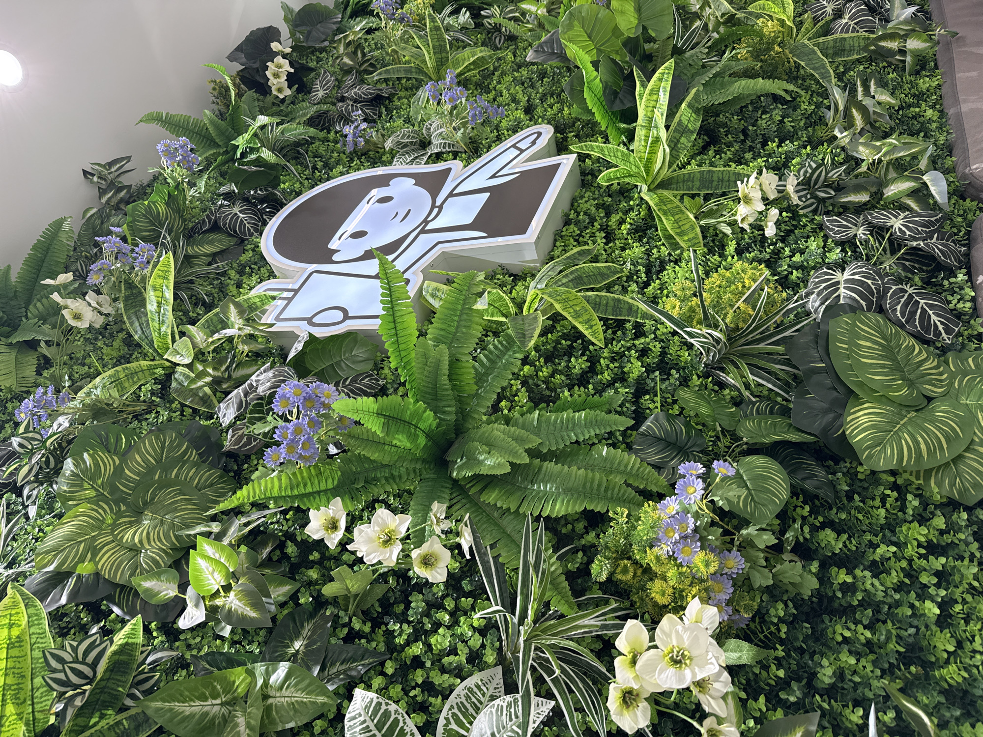 The image shows a wall decorated with various artificial plants and flowers, forming a green wall or vertical garden. A logo with a cartoon-like character is mounted on the wall. The plants include ferns, leafy plants with varied patterns, and small white and purple flowers. The green wall is dense, with the plants arranged closely together. The location is in Krasnoyarsk, Russia.