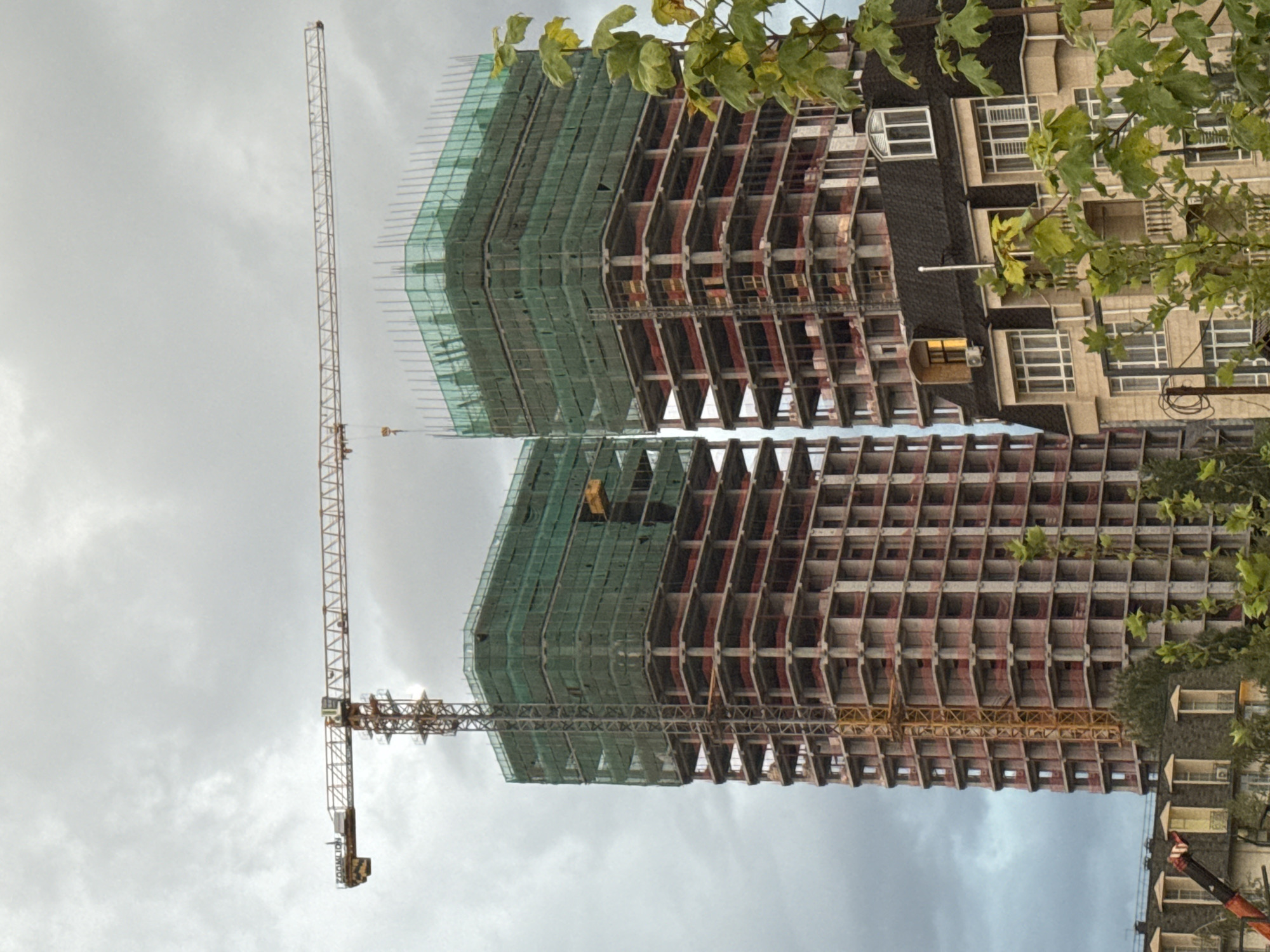 A wide shot captures the construction of two high-rise buildings against a cloudy sky. A yellow tower crane is prominently positioned, extending horizontally over the buildings. The buildings themselves are in various stages of construction, with visible concrete frames and scaffolding covered in green netting. A smaller, more complete building with a dark roof and multiple windows is visible in the foreground, partially obscuring the construction site. Green foliage is present in the lower right corner of the image. The scene is an outdoor urban environment in Bishkek City, Kyrgyzstan.