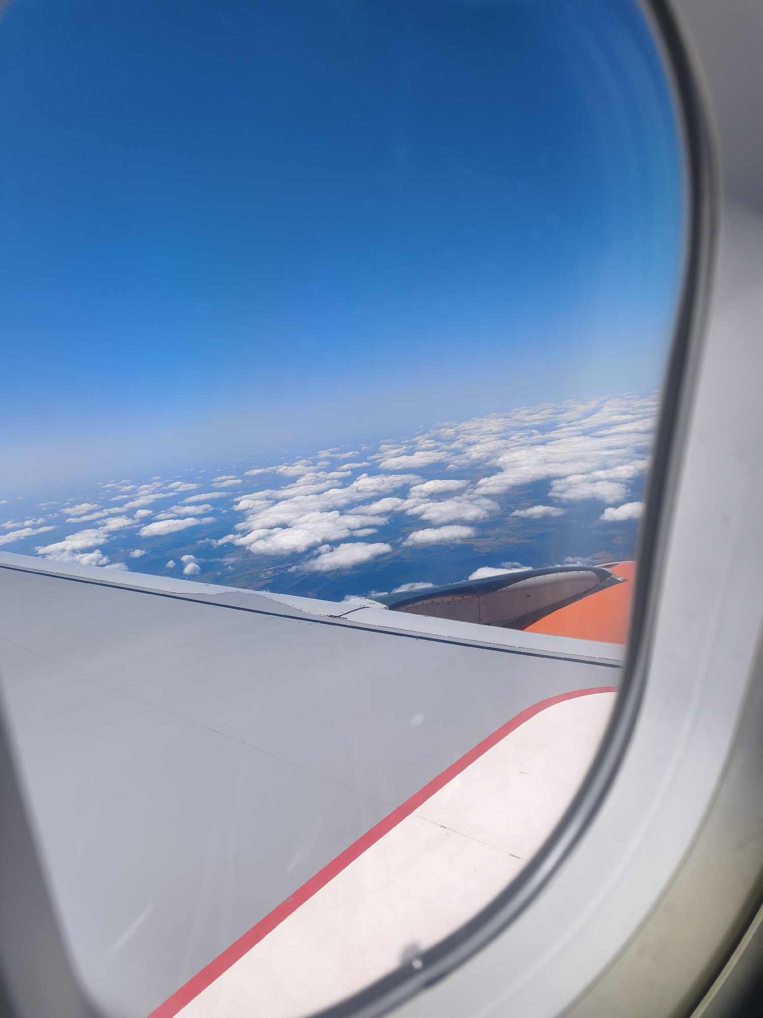 The image is an aerial view from an airplane window, looking out at a blue sky with scattered white clouds. Below the clouds, a landscape of green fields and bodies of water is visible. The wing of the airplane, painted gray with orange accents, occupies the foreground. The scene captures a clear day with good visibility, suggesting good weather conditions for flying. There is no visible indication of the specific location in Calau, Germany.