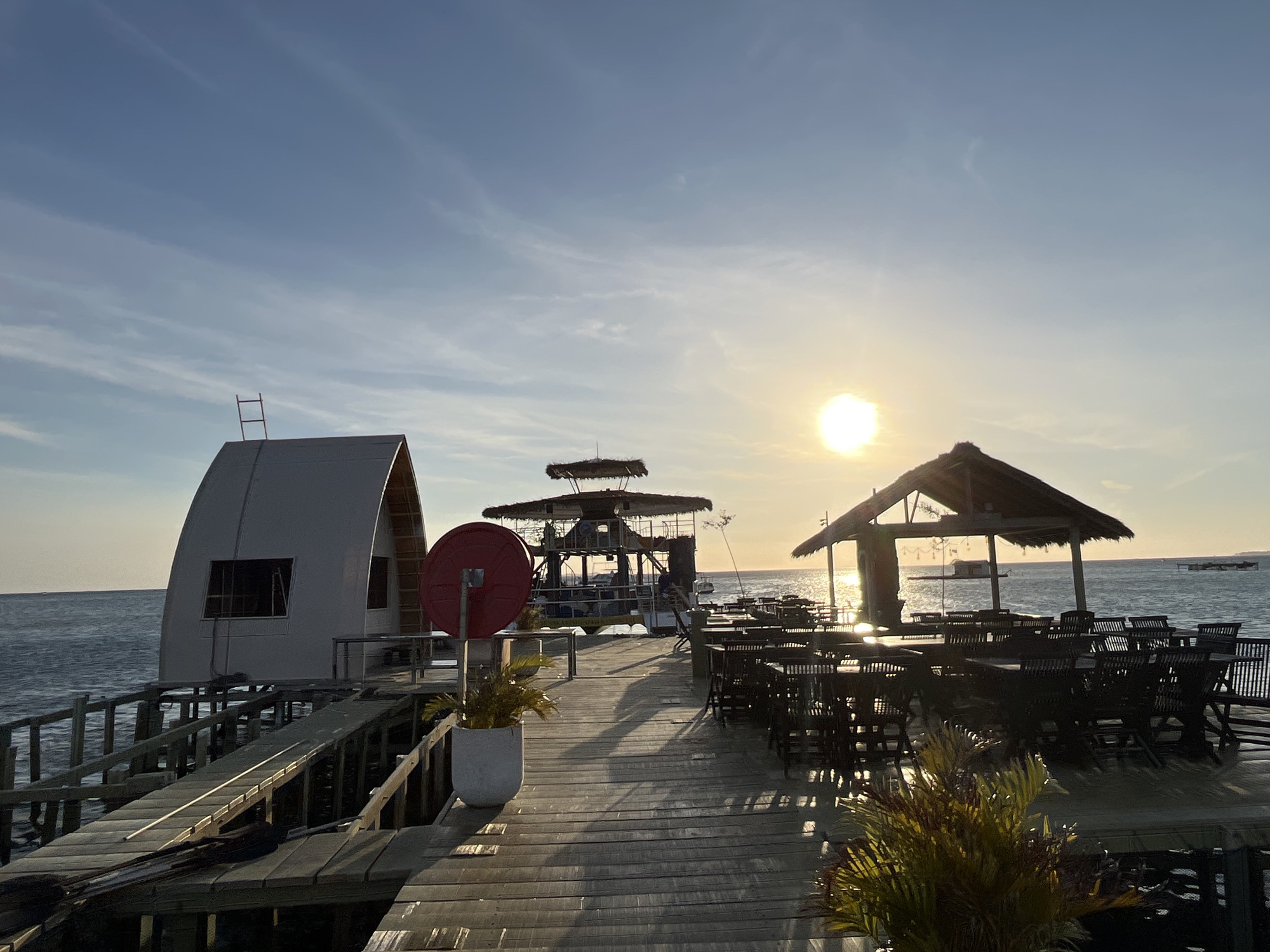 The image shows a wooden pier extending into the sea at Karimunjawa, Indonesia, during a sunset. On the left, there is a white, arced building under construction. To the right, an outdoor dining area with dark wooden tables and chairs is set up under a thatched-roof pavilion. The sun is setting on the horizon, casting a warm glow across the water and the pier, creating long shadows. The sea is calm with a few distant structures or boats visible on the water. The sky is a pale blue with wispy clouds.