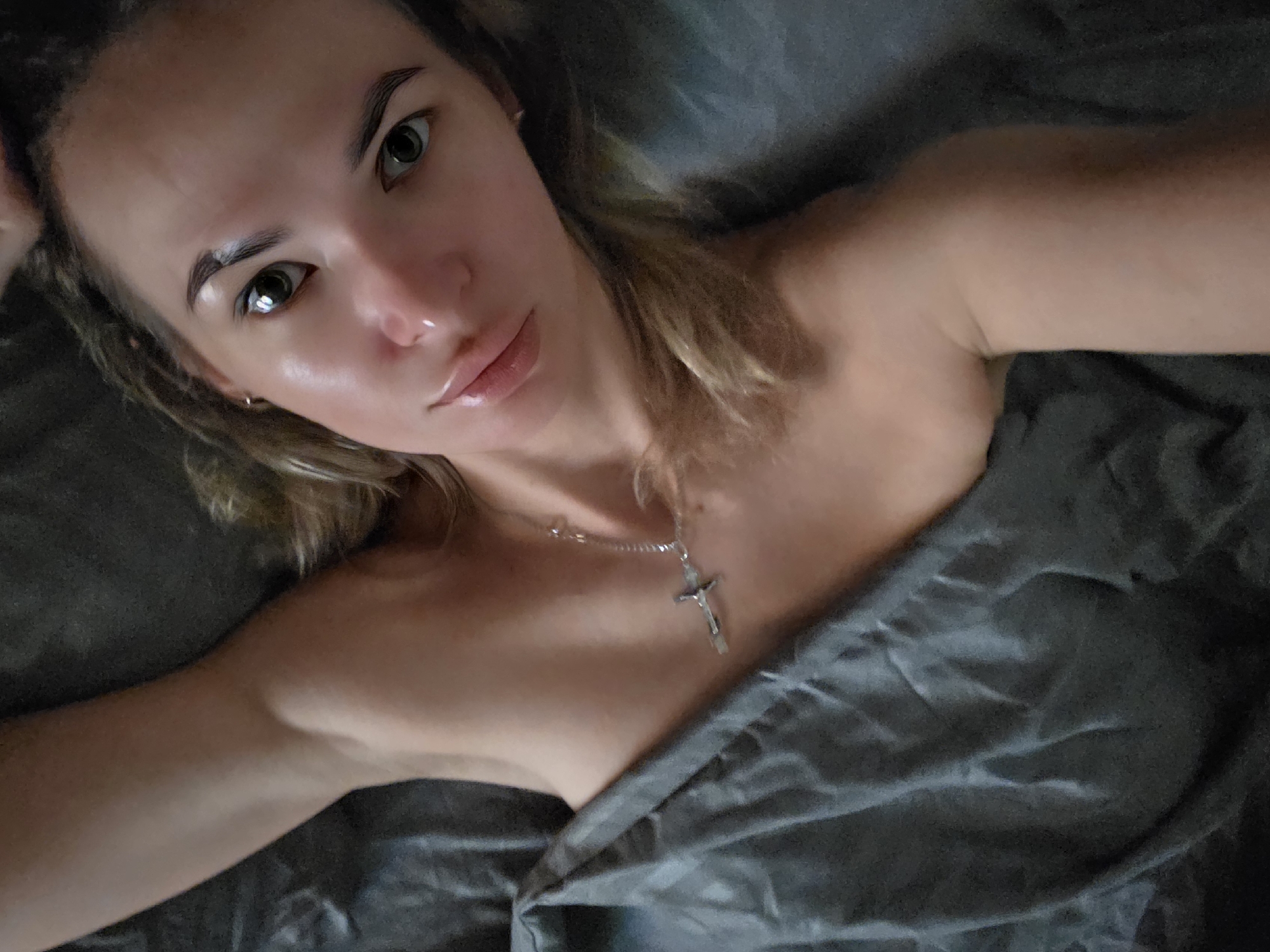 A young adult female is depicted lying in bed, photographed from an overhead perspective. Her head is positioned towards the upper right of the frame, with her body extending diagonally downwards to the left. She is looking directly at the camera. Her complexion appears fair with a slight sheen on her forehead, nose, and lips. Her eyes are dark, and her eyebrows are dark and well-defined. She has full lips and what appears to be a small stud piercing in her right nostril. Her hair is light brown or blonde, somewhat disheveled, and spread across the dark grey bedding behind her head.
Her upper torso, including her right shoulder and arm extending upwards out of frame, appears bare. A silver-colored chain necklace with a small crucifix pendant hangs around her neck, resting on her chest. A small, simple stud earring is visible in her left ear. The lower portion of her body is obscured by a rumpled dark grey sheet or duvet, which covers her from approximately mid-chest downwards. The immediate environment consists solely of this dark grey fabric, offering no additional contextual details.
The visual information in the image does not permit the identification of a specific location within the city of Chaikovsky, Russia.