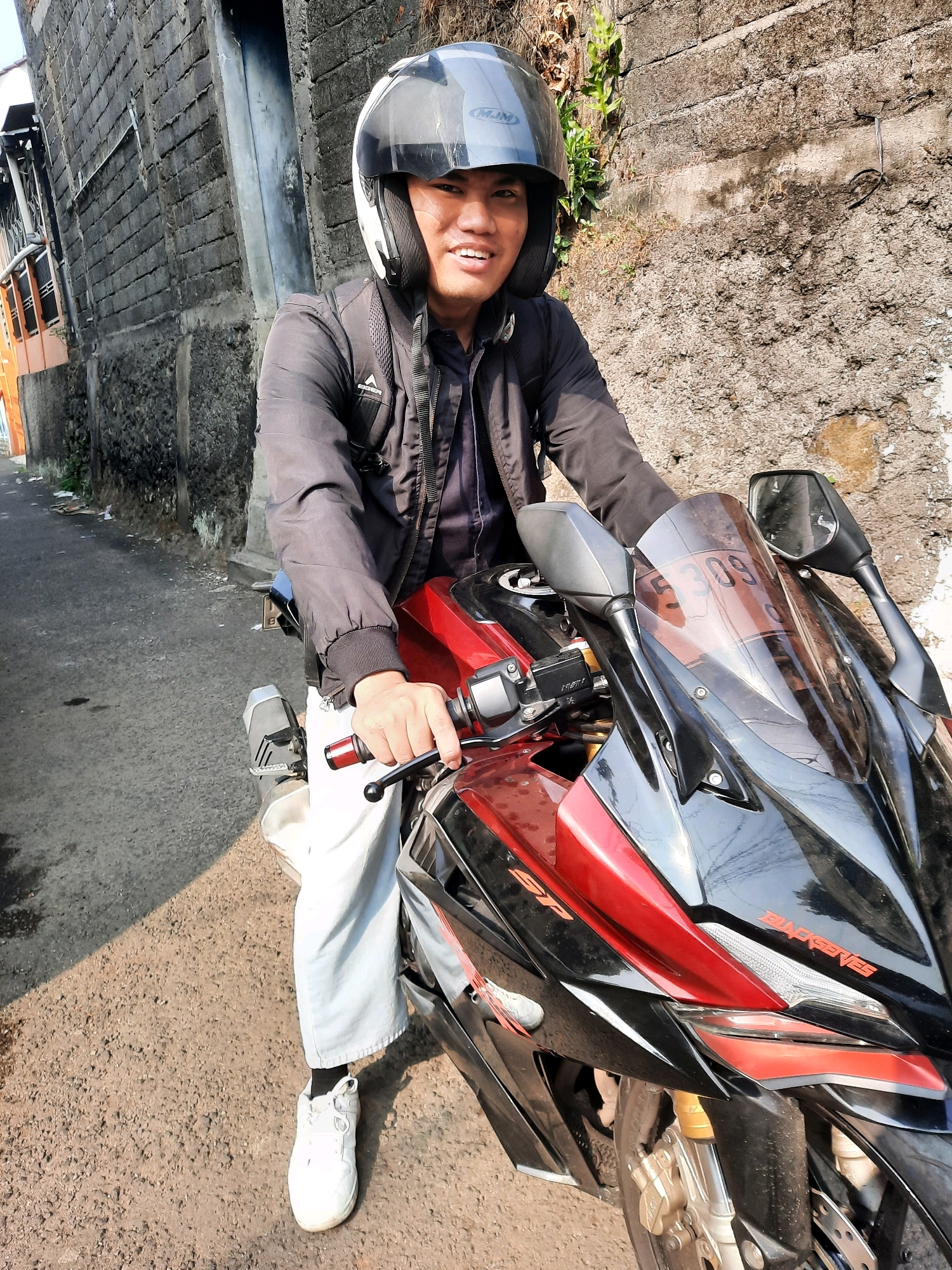 A man wearing a black jacket, white pants, and a white helmet is sitting on a red and black motorcycle. He is looking towards the camera and smiling. The motorcycle has a black windshield and two mirrors. The man's left hand is on the handlebars. The background shows a textured stone wall, a narrow street, and some greenery. The scene appears to be in an urban environment. The license plate of the motorcycle has the number "5309".
