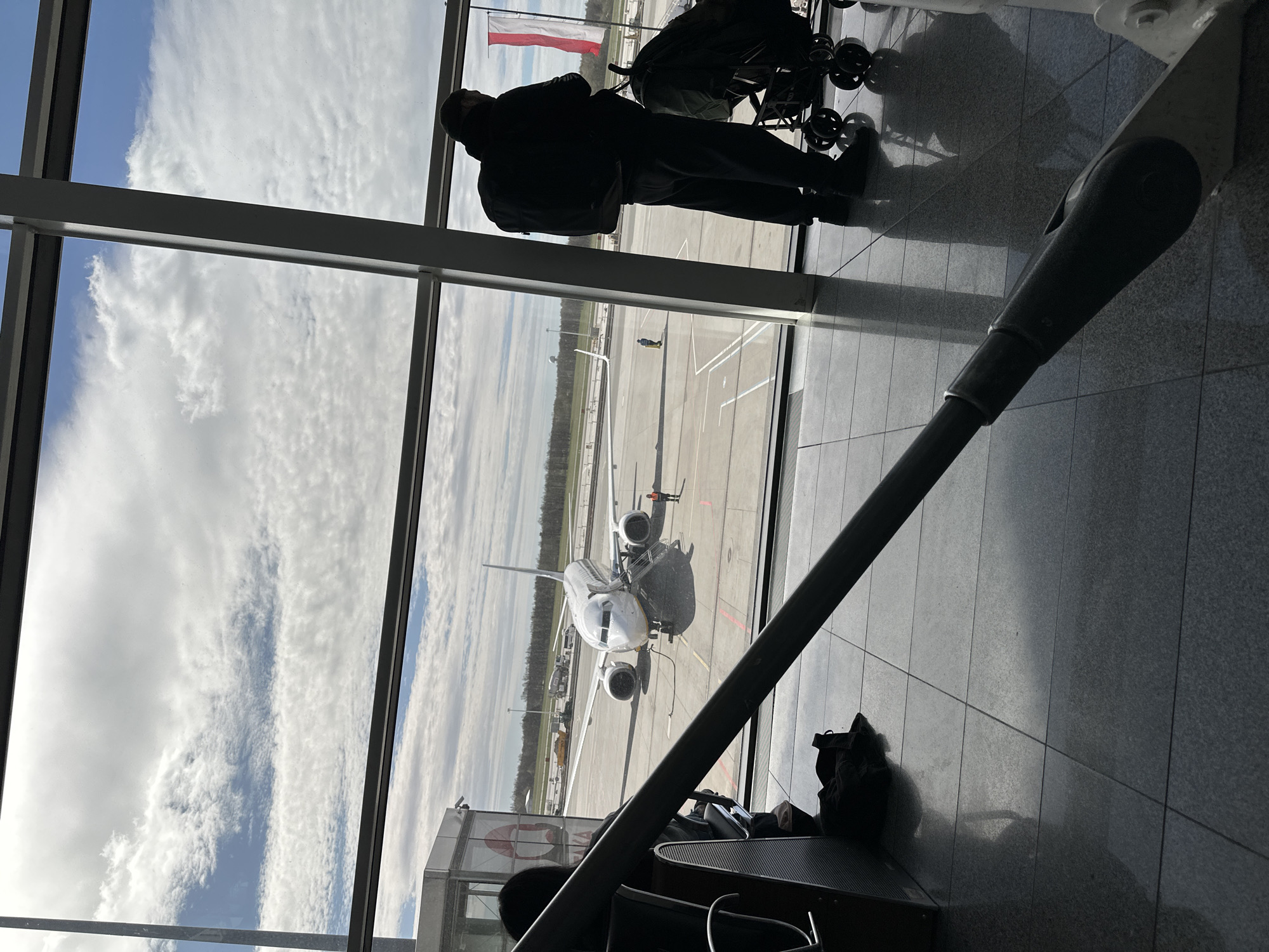 The image depicts an airport scene, potentially in Wrocław, Poland. A person stands at a large window, silhouetted against the bright daylight outside. Next to the person is a stroller. Through the window, an airplane is visible on the tarmac. In the distance, a Polish flag flies. The interior shows airport seating, with personal belongings (bags) on the floor. The structure holding up the building's wall in which the large windows are inserted is visible as well.