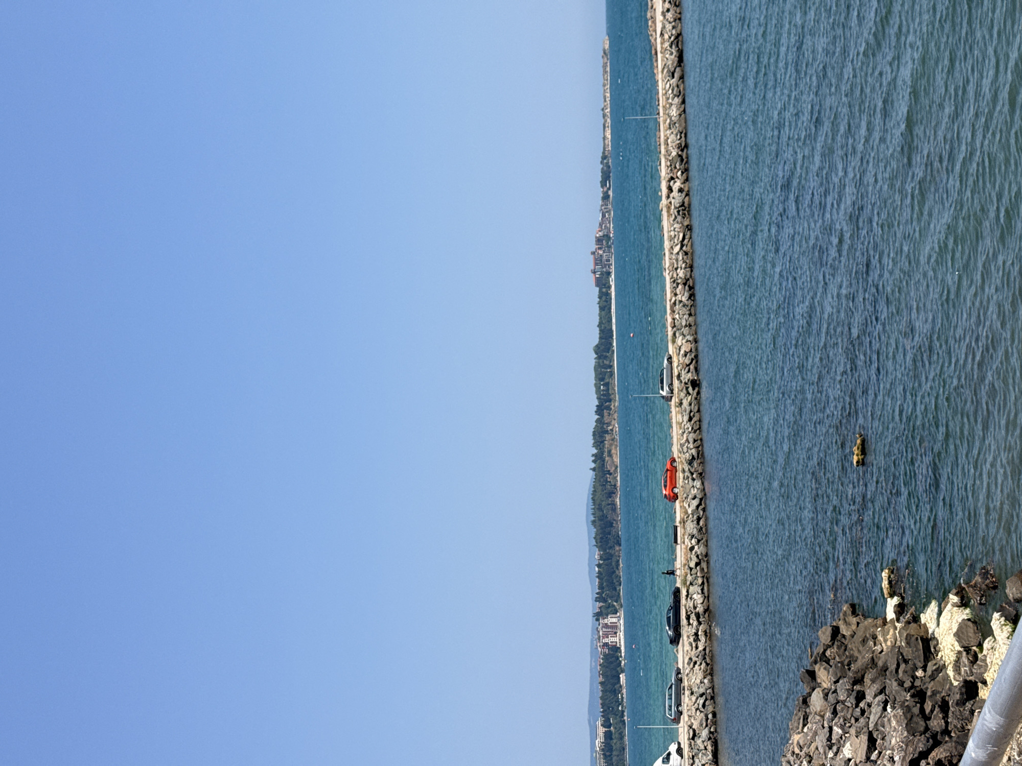 The image captures a scenic view of a pier in Ravda, Bulgaria, extending into a clear blue sea under a cloudless sky. The pier, constructed from large rocks, is lined with a paved walkway. Several vehicles, including a white van, a dark-colored sedan, a black sports car, and a red car, are parked or driving along the walkway. A lone pedestrian is visible walking on the pier. In the distance, the coastline of Ravda is dotted with buildings, including hotels and residential structures, surrounded by lush green trees. The sea is calm, with gentle ripples on its surface. In the foreground, a rocky embankment leads down to the water's edge.