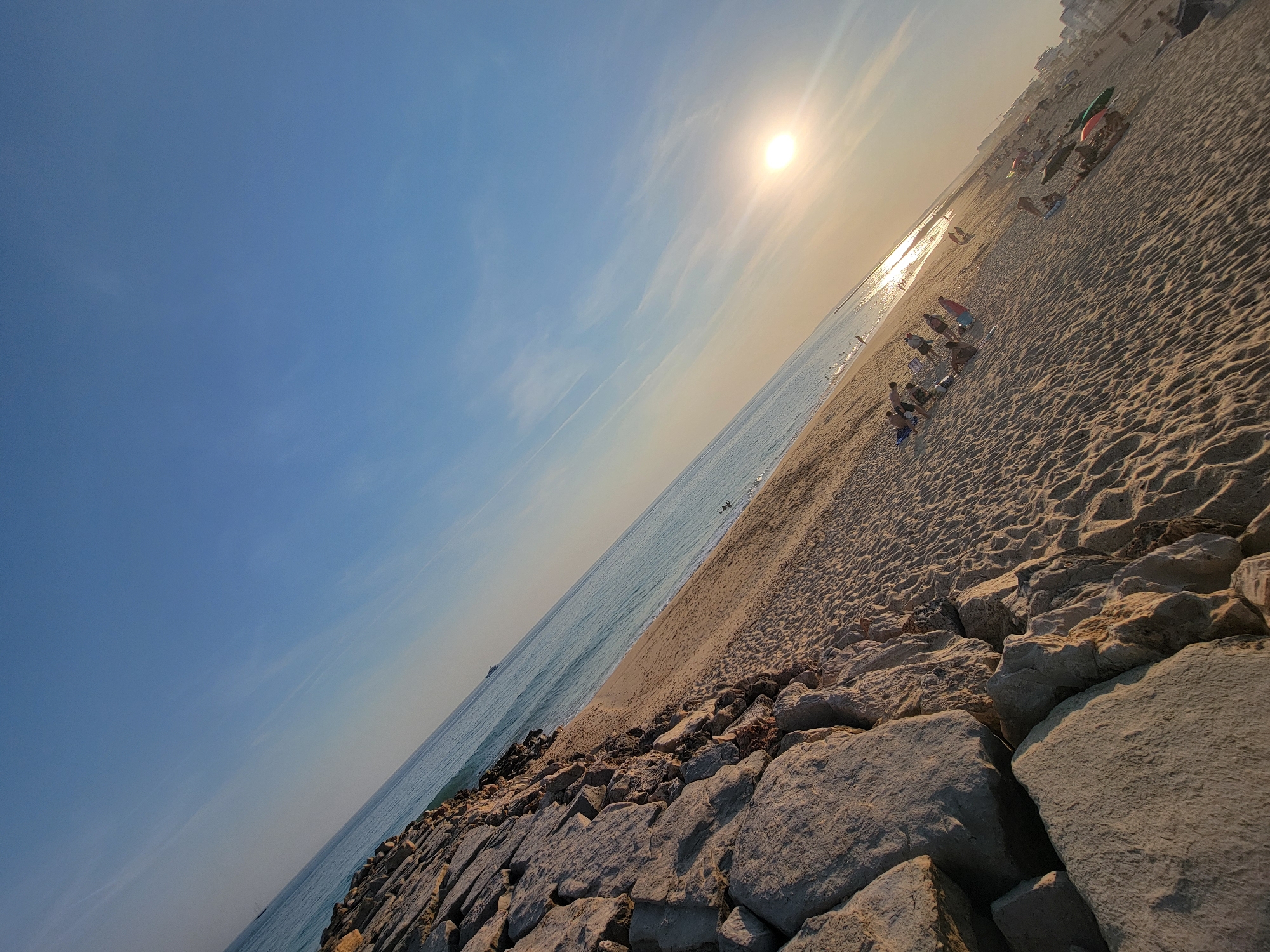 The image captures a wide, sandy beach in Quarteira, Portugal, bathed in the warm glow of the setting sun. The perspective is tilted, with a large, rocky seawall dominating the left foreground, its coarse texture and varying shades of grey and brown sharply defined. Beyond the seawall, the calm, turquoise ocean stretches towards the horizon, reflecting the sunlight.
The beach itself is populated with a scattering of people enjoying the late afternoon. Several individuals and small groups are visible, some lounging on towels or beach chairs, others standing or walking along the shoreline. Umbrellas dot the sand, providing patches of shade. In the distance, the coastline is lined with buildings, suggesting an urban setting. The sand is textured, with ripples and footprints indicating recent activity. The sky above is a clear, gradient blue, transitioning to a lighter hue near the horizon where the sun, a bright, intense orb, casts a long, shimmering reflection across the water. A few wispy clouds streak across the sky. The overall impression is one of a peaceful, late summer afternoon at a popular seaside resort.