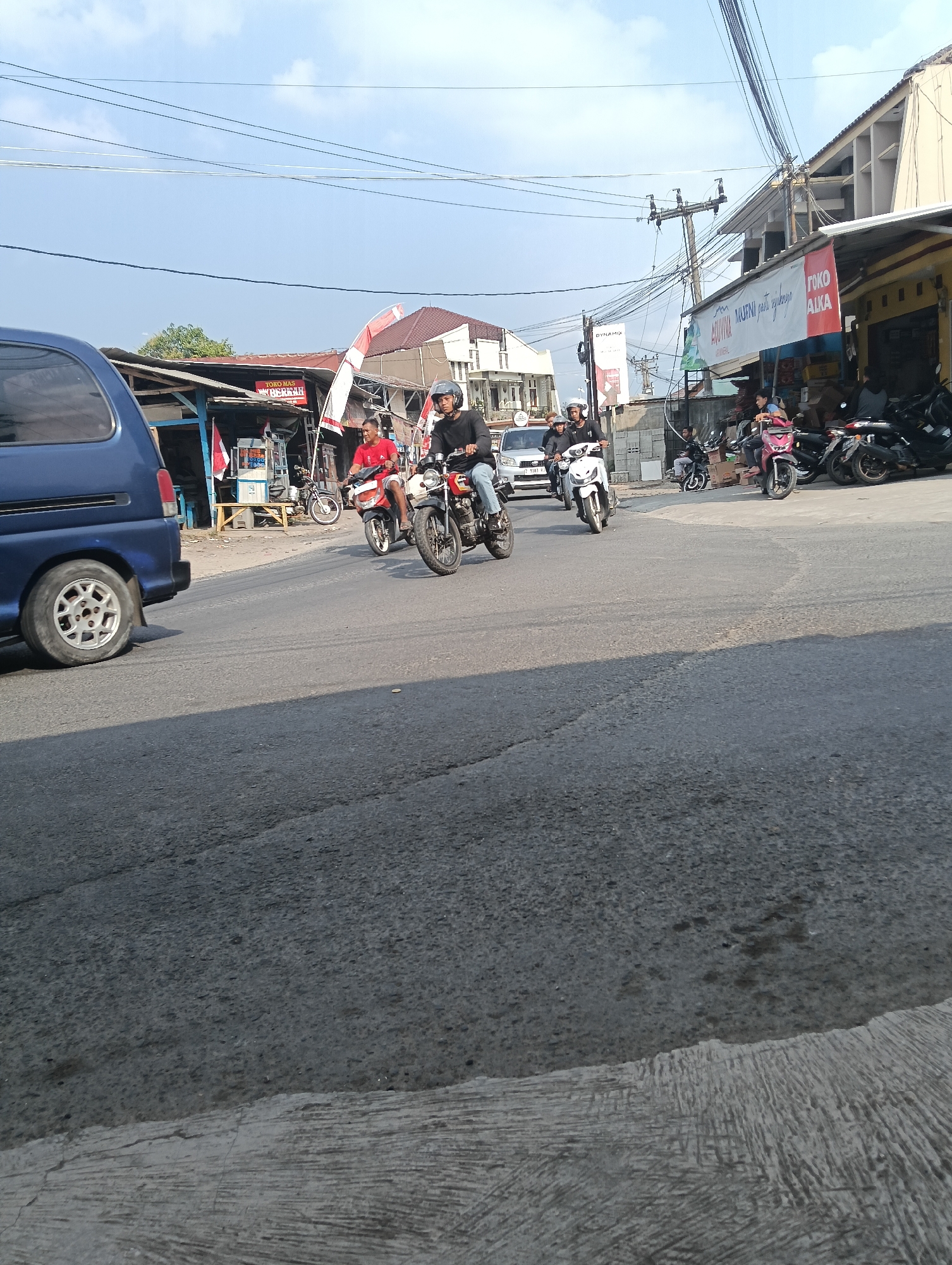 In Puspahiang, Indonesia, on a bright, clear day, a street scene unfolds. A blue van is parked on the left side of the road, facing away from the camera. The road is paved with asphalt and shows signs of wear. On the right side of the road, a commercial area is visible, with several shops and buildings. One shop has a yellow facade and a red and white sign that reads "TOKO ALKA". Another shop has a white banner with Indonesian text, likely advertising.
Several motorcycles are on the road, indicating traffic. Two men on motorcycles are prominent in the mid-ground. One is wearing a red shirt and a helmet, riding a red and black motorcycle. The other, in a black shirt and helmet, is riding a black motorcycle with some chrome accents. Both are moving away from the camera. Further back, more motorcycles and riders are visible, some stopped and some moving. There are also parked motorcycles in front of the shops on the right.
The background features a blue sky with scattered white clouds. Power lines crisscross the sky, supported by utility poles. Buildings of varying architectural styles line the street, some appearing to be shops or small businesses. The overall impression is a typical bustling street scene in a commercial area of a town in Indonesia.